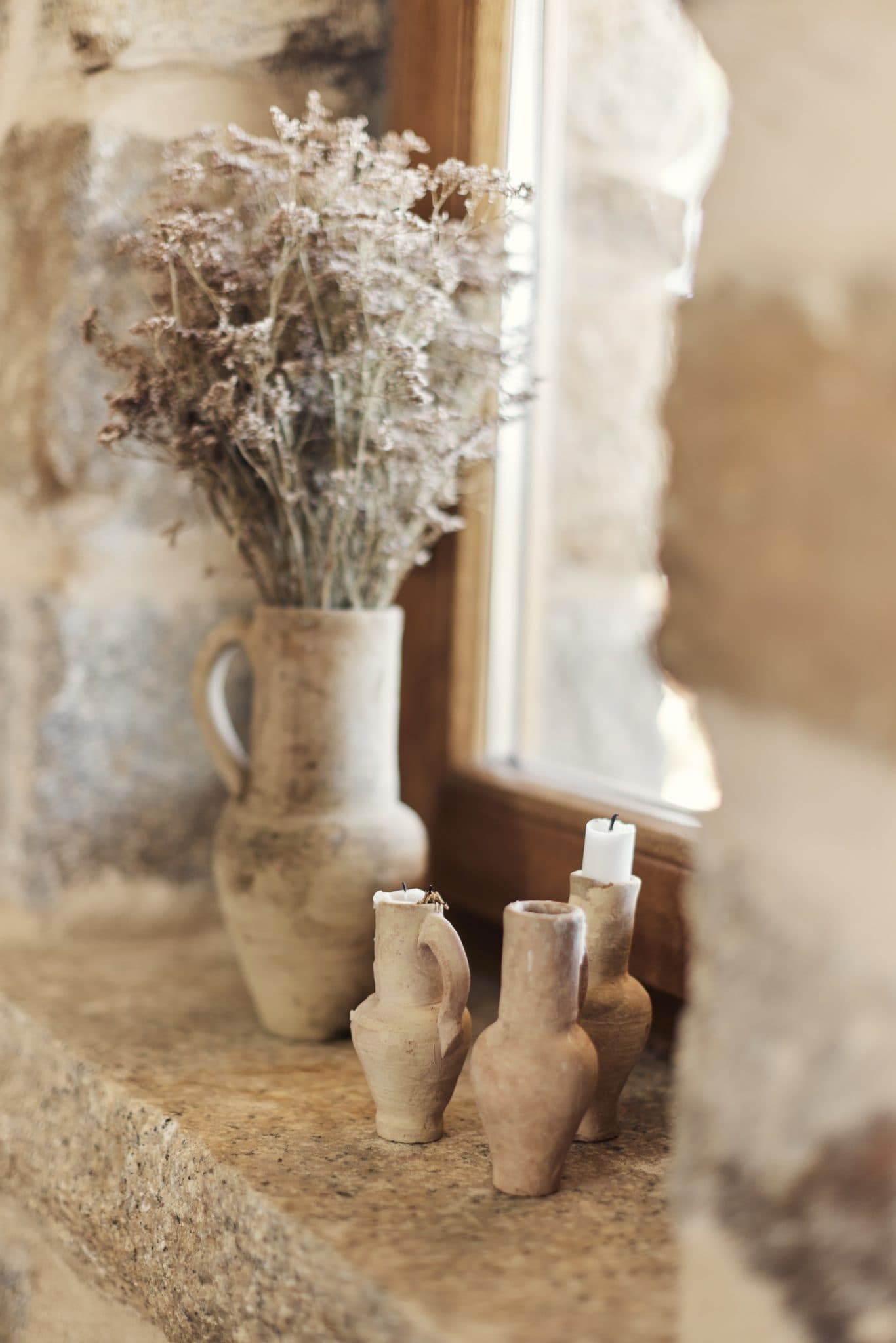 A rustic scene featuring a stone windowsill with a beige ceramic vase holding dried flowers. In the foreground, three small beige ceramic vases each contain white candles. The window and stone wall enhance the warm and natural atmosphere.
