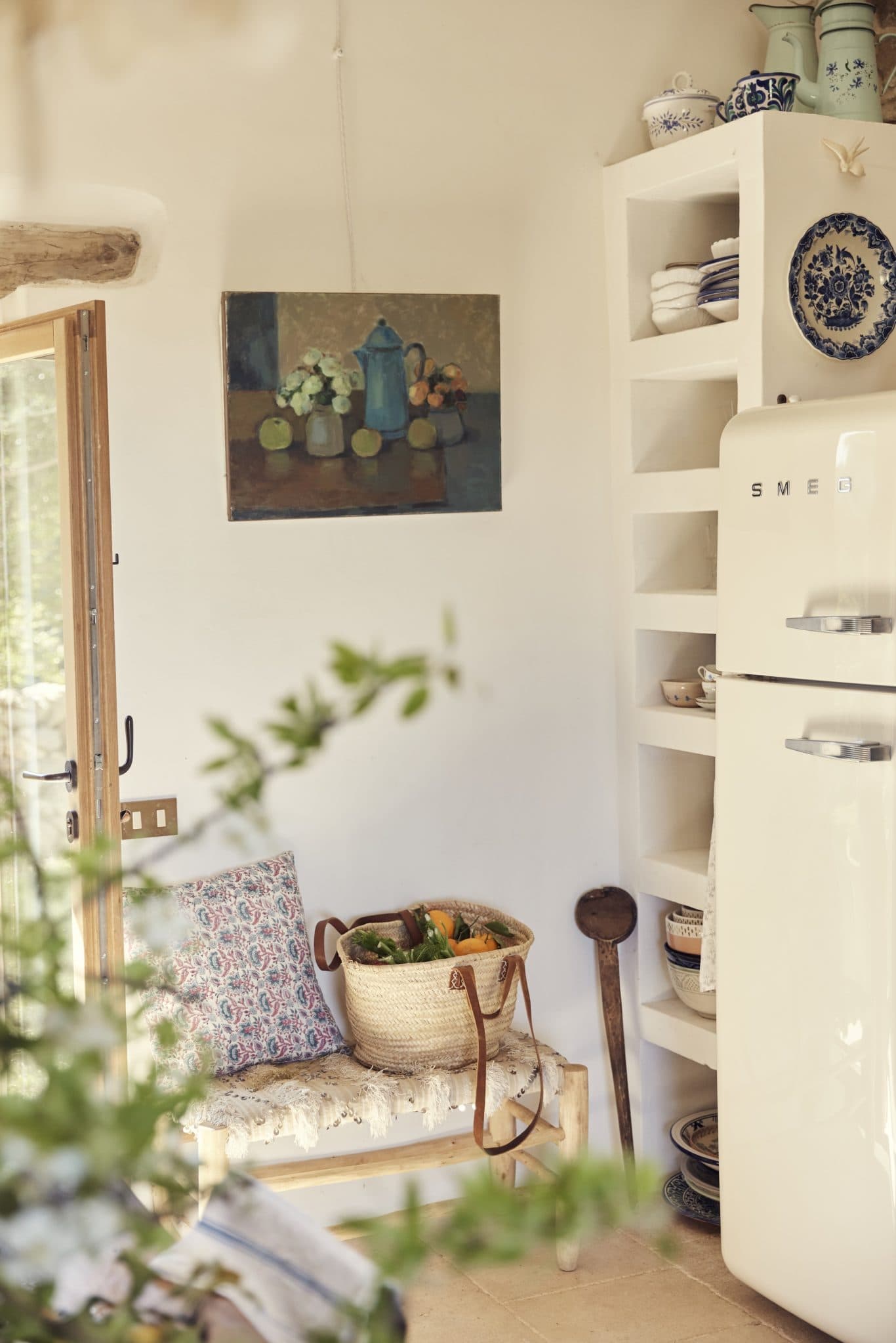 A cozy kitchen corner with a vintage refrigerator, white open shelves holding various dishes, and a bench with a floral cushion and a woven basket filled with fresh produce. A still life painting of fruits and a pitcher hangs on the wall above the bench.