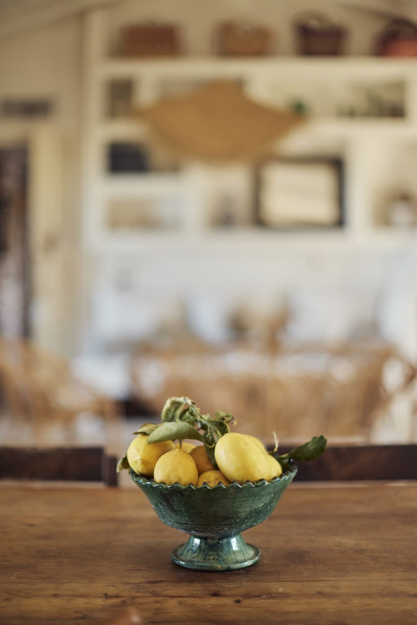 A green ceramic bowl filled with yellow lemons sits on a wooden table. The softly lit, blurred kitchen in the background, with various items on the shelves, evokes a warm and rustic atmosphere.
