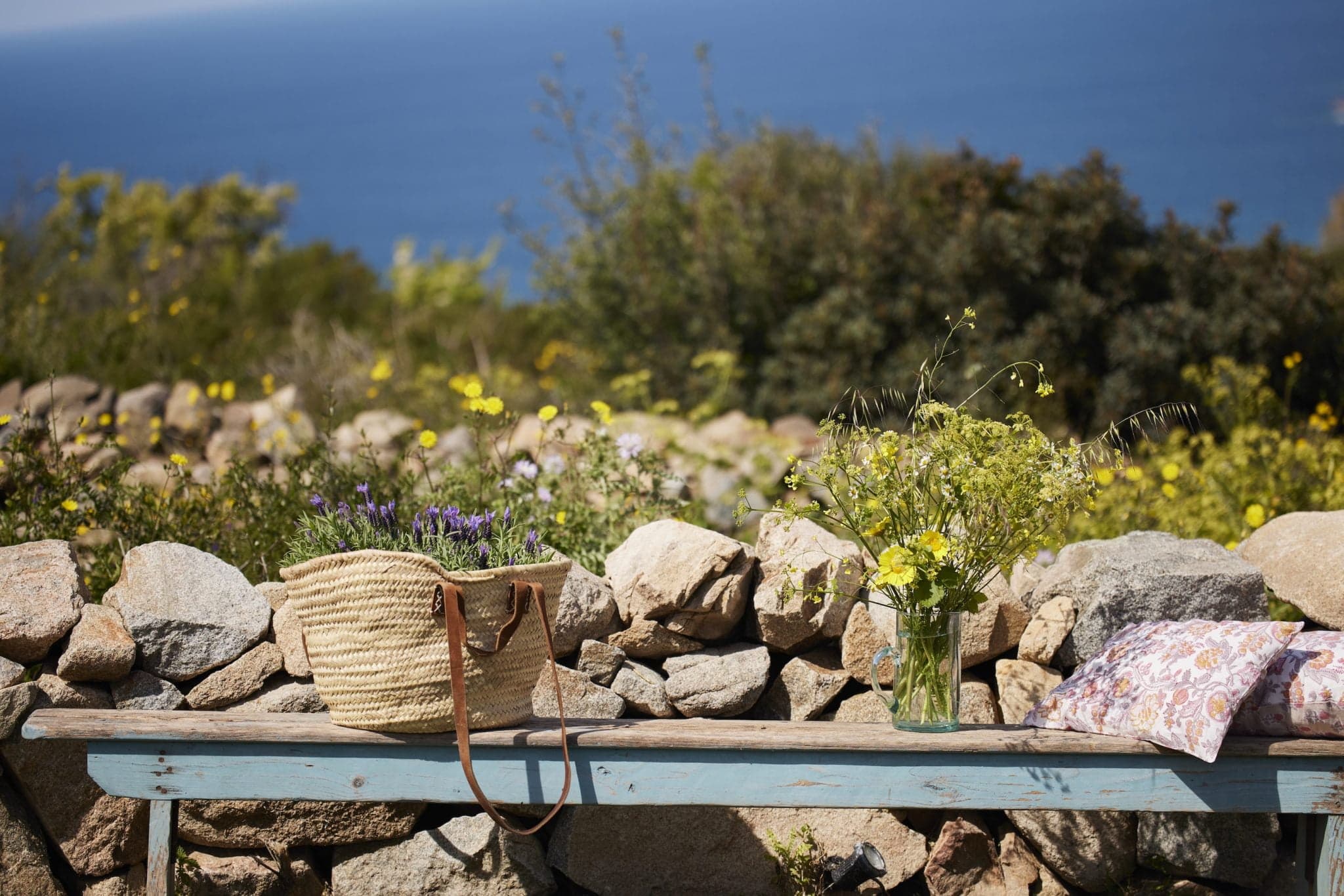 A rustic wooden bench, set against a stone wall, overlooks a picturesque coastal landscape. On the bench sits a straw bag filled with flowers, a glass vase holding yellow blossoms, and a floral-patterned cushion, with the blue sea visible in the background.