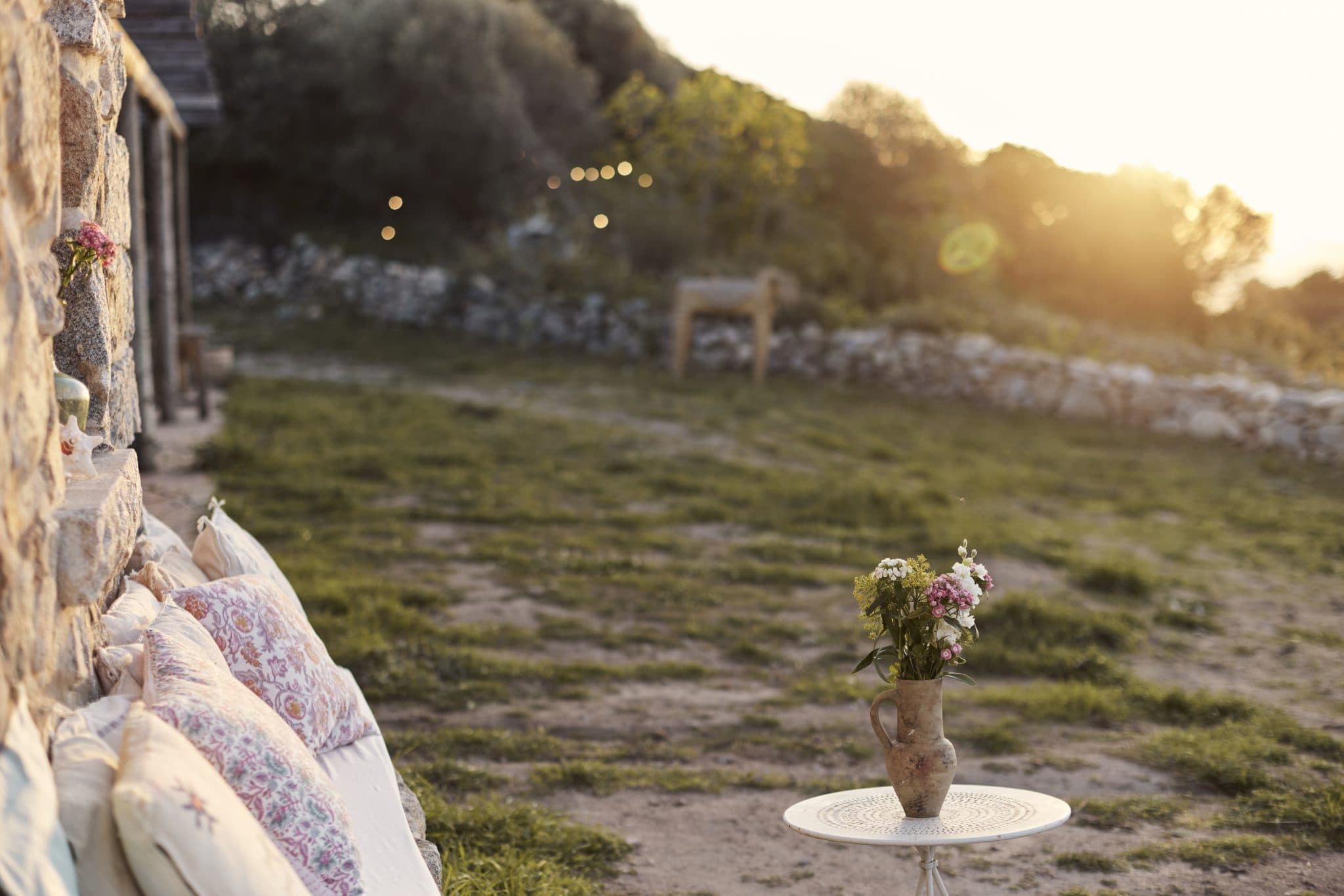 A sunny outdoor scene with a rustic vibe, featuring a stone wall adorned with colorful cushions on the left. In the foreground, a round table holds a vase with flowers. The background showcases greenery with stone walls and the soft, warm light of the setting sun.