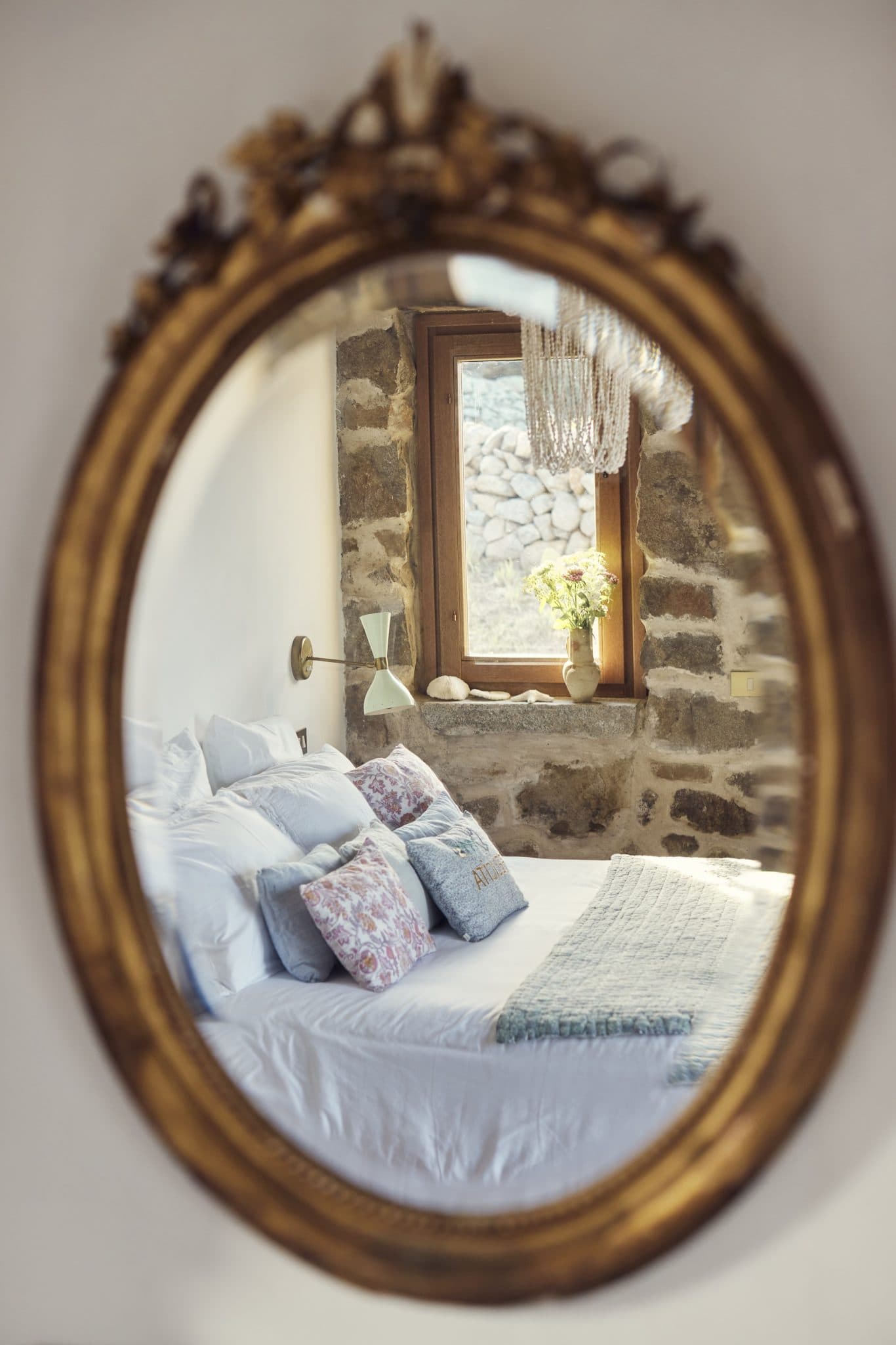 A cozy bedroom is visible through an ornate oval mirror. The room features stone walls, a bed with white linens and decorative cushions in various colors. A wooden-framed window looks out onto an exterior stone wall, and a vase with flowers rests on a bedside table.