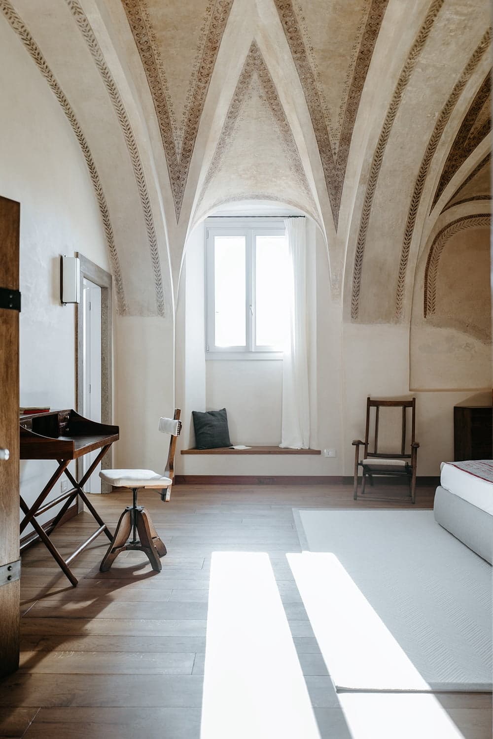 Une pièce spacieuse avec de hauts plafonds voûtés et une fenêtre au-dessus d'un banc intégré. La pièce comprend deux chaises, un bureau en bois et une décoration minimaliste, créant une atmosphère sereine et aérée. La lumière du soleil pénètre par la fenêtre, illuminant le plancher en bois.