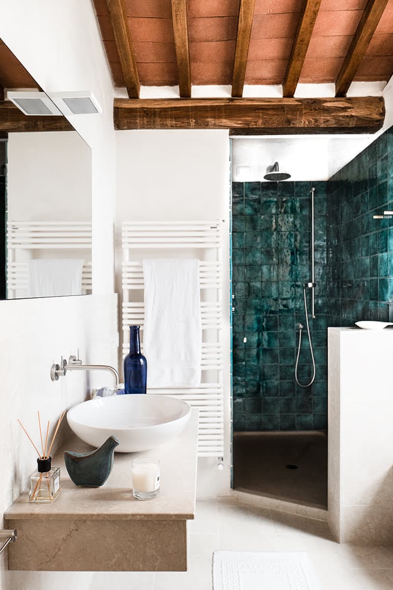 Une salle de bains moderne comprend un lavabo avec un plan de travail orné d'un vase bleu, d'une bougie et d'un diffuseur à roseaux. Une serviette blanche est suspendue à un radiateur et la douche, à l'arrière-plan, présente des murs carrelés bleu-vert avec une porte en verre. Le plafond présente des poutres en bois apparentes.