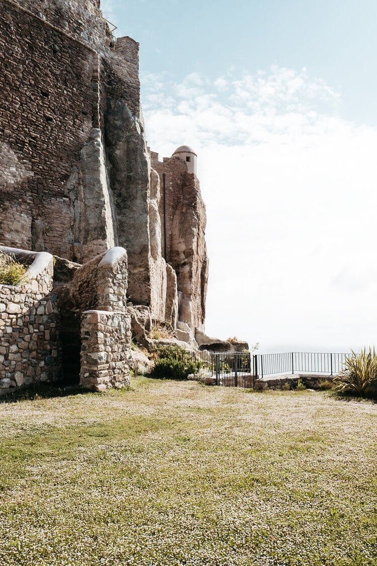 Une forteresse médiévale en pierre se dresse sur une falaise rocheuse, avec des murs érodés par les intempéries et une petite tourelle visible. La forteresse surplombe une zone herbeuse qui mène à une clôture métallique, au-delà de laquelle le ciel rencontre l'horizon. Le soleil projette une douce lumière sur la scène.