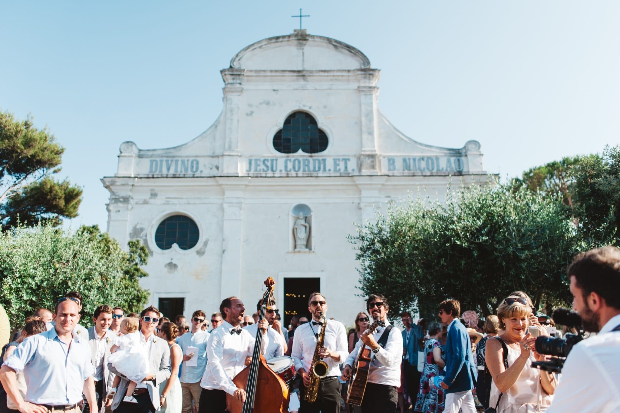 Une foule animée, certains tenant des instruments de musique, se rassemble devant une église blanche surmontée d'une croix. L'église est dotée de fenêtres en arc et d'une inscription en latin. Les participants, vêtus de tenues d'été, semblent participer à un événement festif sous un ciel bleu clair.