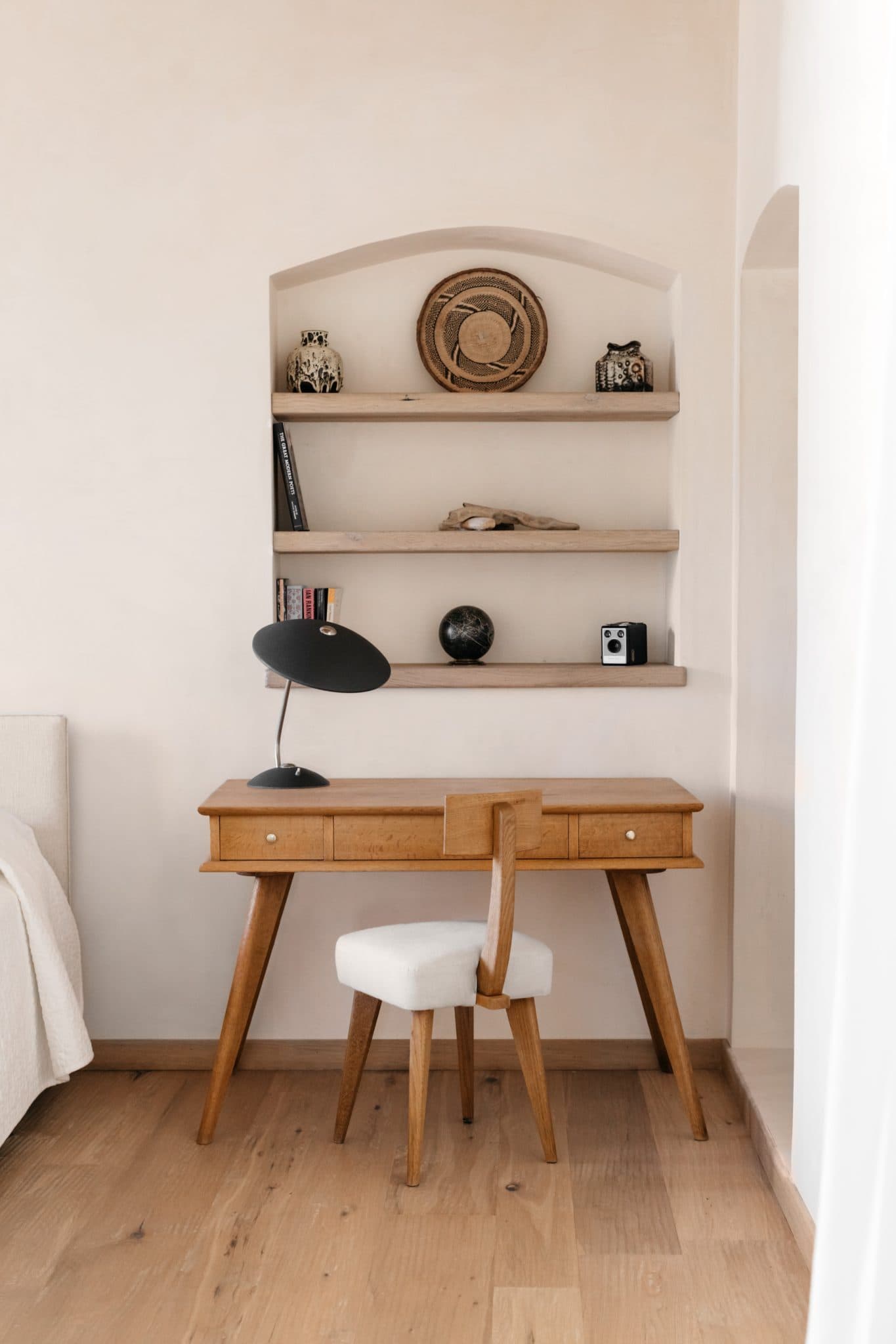 Un bureau à domicile minimaliste comprend un bureau en bois avec une lampe noire moderne et une chaise rembourrée. Au-dessus du bureau se trouvent des étagères en bois intégrées contenant des livres, des bols décoratifs et un appareil photo vintage. La pièce a des murs de couleur claire et du parquet.