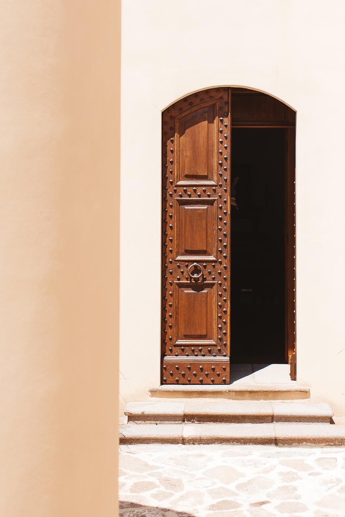 Une porte en bois légèrement ouverte et décorée est encastrée dans un mur en pierre beige, ce qui permet d'apercevoir l'intérieur sombre. La porte présente des motifs complexes, notamment des clous le long de ses bords. Des marches en pierre mènent à la porte et la lumière du soleil projette des ombres sur le sol.