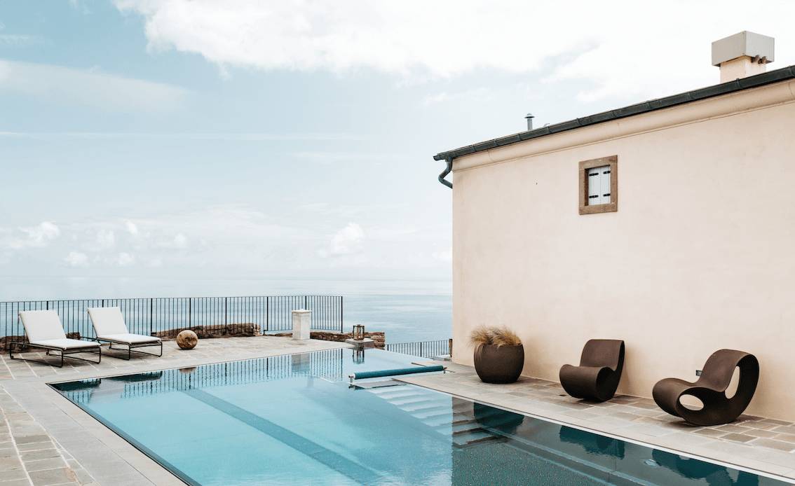 Un espace piscine extérieur moderne avec deux chaises longues blanches, deux chaises élégantes marron foncé et une plante en pot. Le bord de la piscine donne sur une vue paisible sur l'océan, reflétant un ciel bleu clair avec un minimum de nuages. Un bâtiment minimaliste avec une petite fenêtre est adjacent à la piscine.