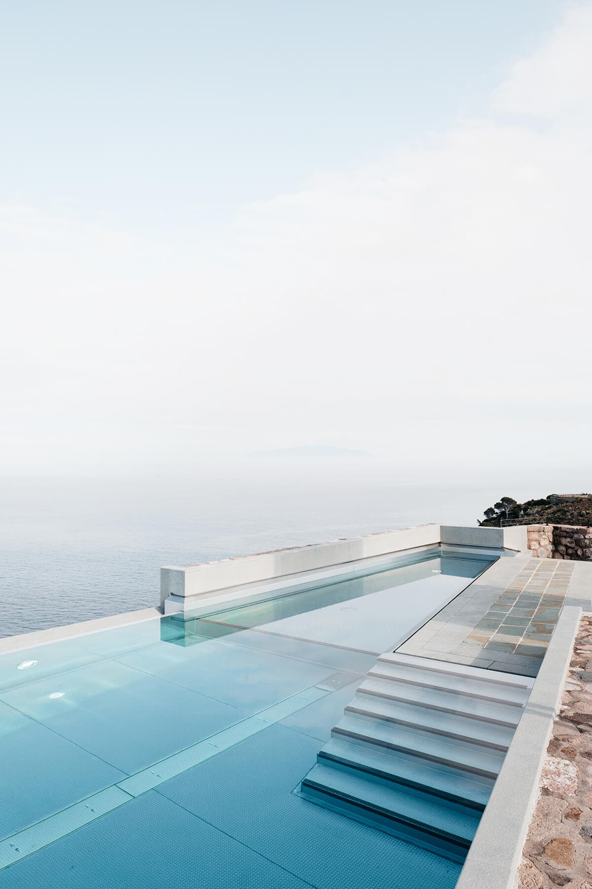 Une piscine à débordement sereine avec une eau bleue claire surplombe un vaste océan calme sous un ciel brumeux. La piscine est dotée de marches immergées et d'un design élégant et moderne, bordé de pierre et de béton. Le cadre tranquille crée une atmosphère paisible et luxueuse.