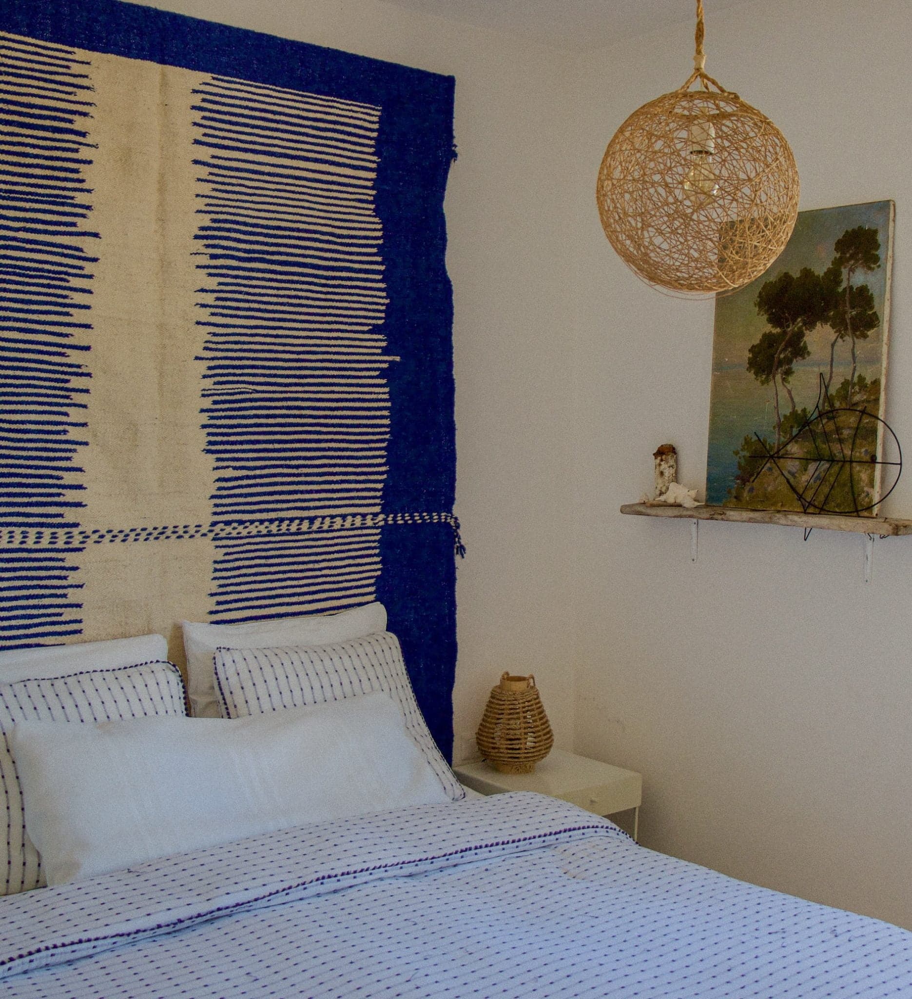 Une chambre douillette est dotée d'un lit avec une literie blanche et bleue, contre un mur orné d'une tapisserie bleue et beige. Une suspension en osier est suspendue au plafond. Une étagère rustique abrite un tableau de paysage et des objets décoratifs, créant une atmosphère chaleureuse et accueillante.