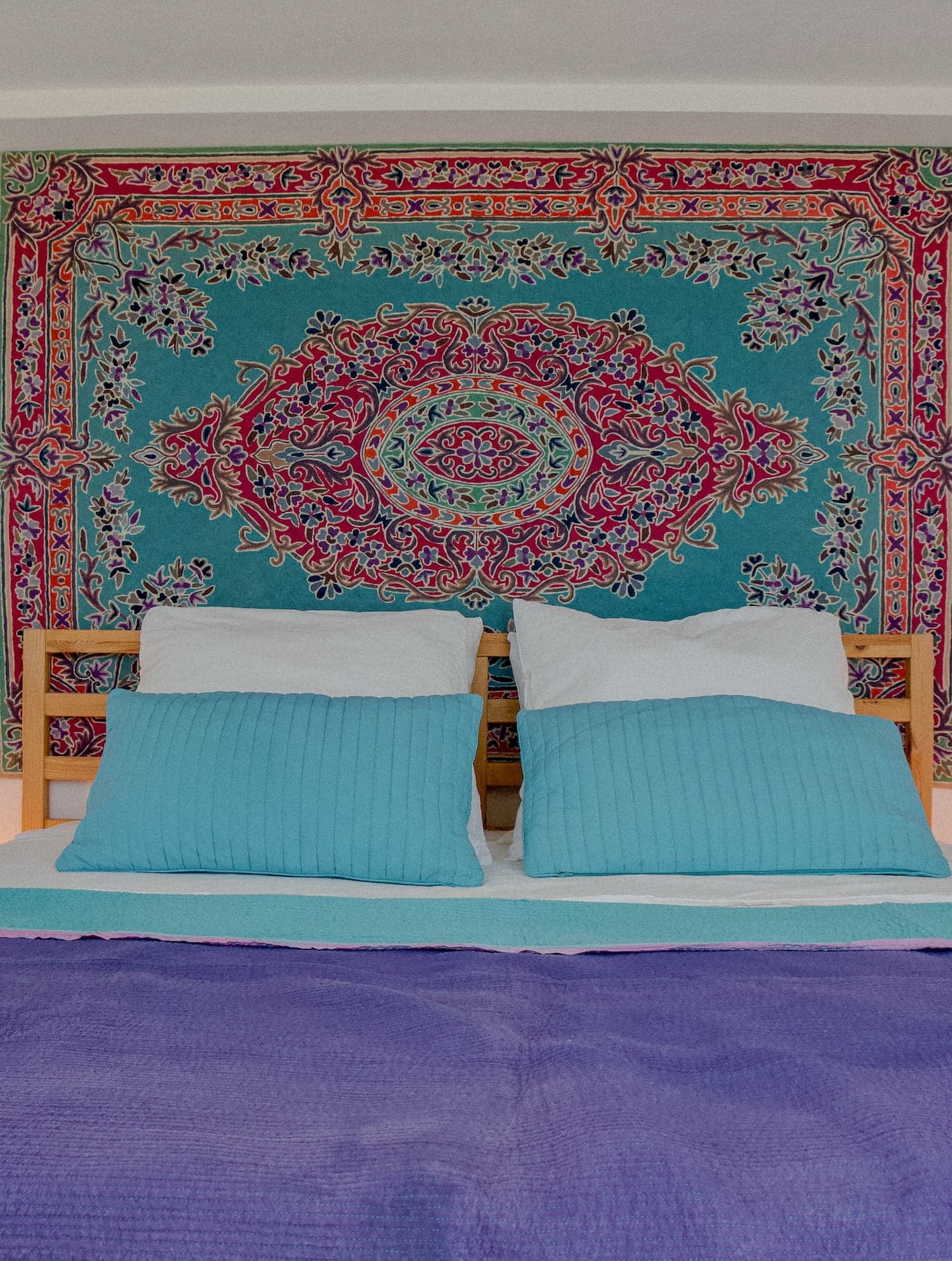 Un lit bien fait avec des oreillers turquoise et une couverture violette est placé contre un mur orné d'une tapisserie complexe et colorée présentant des motifs floraux et géométriques dans des tons de rouge, bleu et violet.