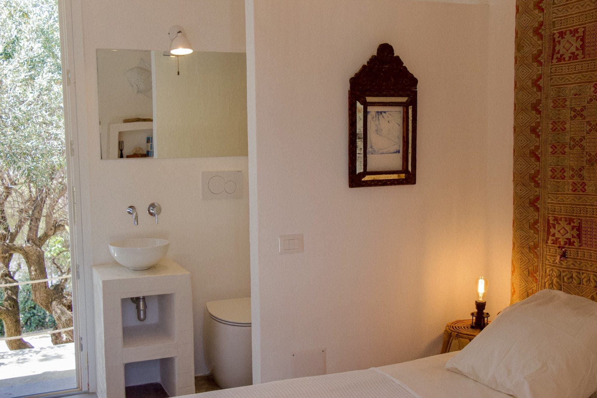 Une chambre confortable et minimaliste avec un mur blanchi à la chaux, un petit lavabo et un miroir au-dessus à gauche. Un tableau encadré et une tapisserie sont fixés sur le mur de droite. Une partie d'un lit soigneusement fait est visible, ainsi qu'une petite table d'appoint sur laquelle repose une lampe allumée.