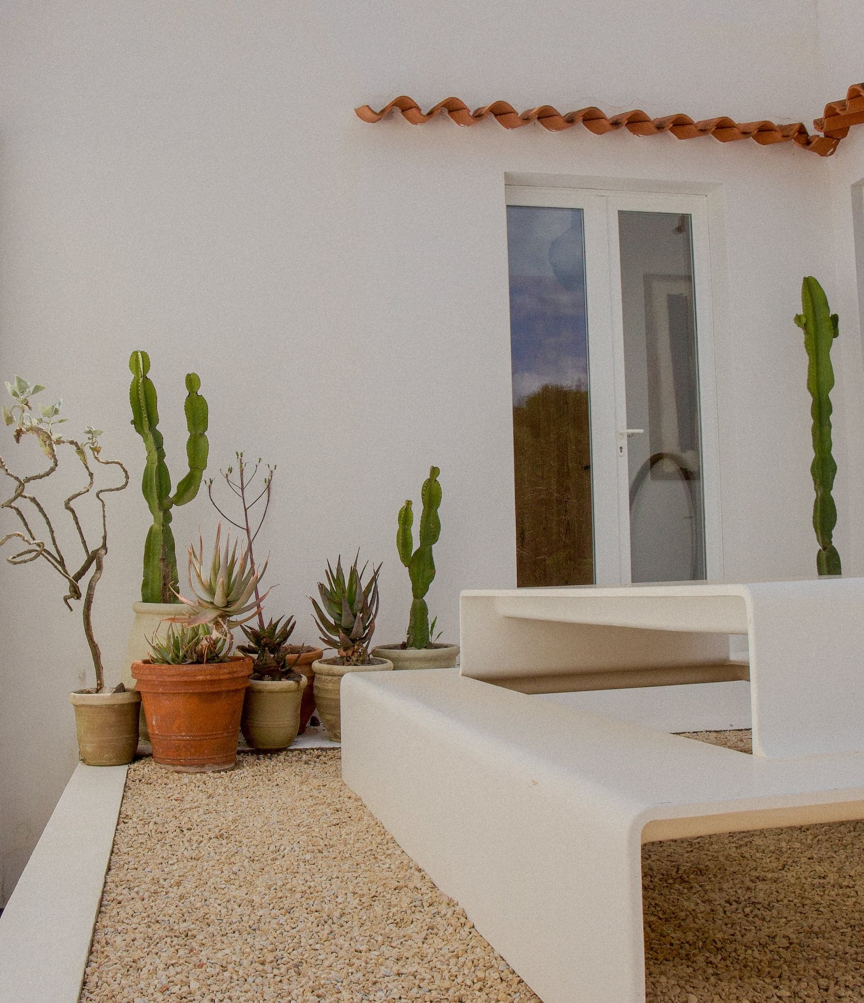 Un espace extérieur minimaliste avec un banc moderne blanc posé sur une surface en gravier. Diverses plantes en pot, dont des cactus et des plantes succulentes, bordent le mur du bâtiment blanc. Une porte avec des panneaux de verre et des tuiles en terre cuite se trouve à l'arrière-plan.