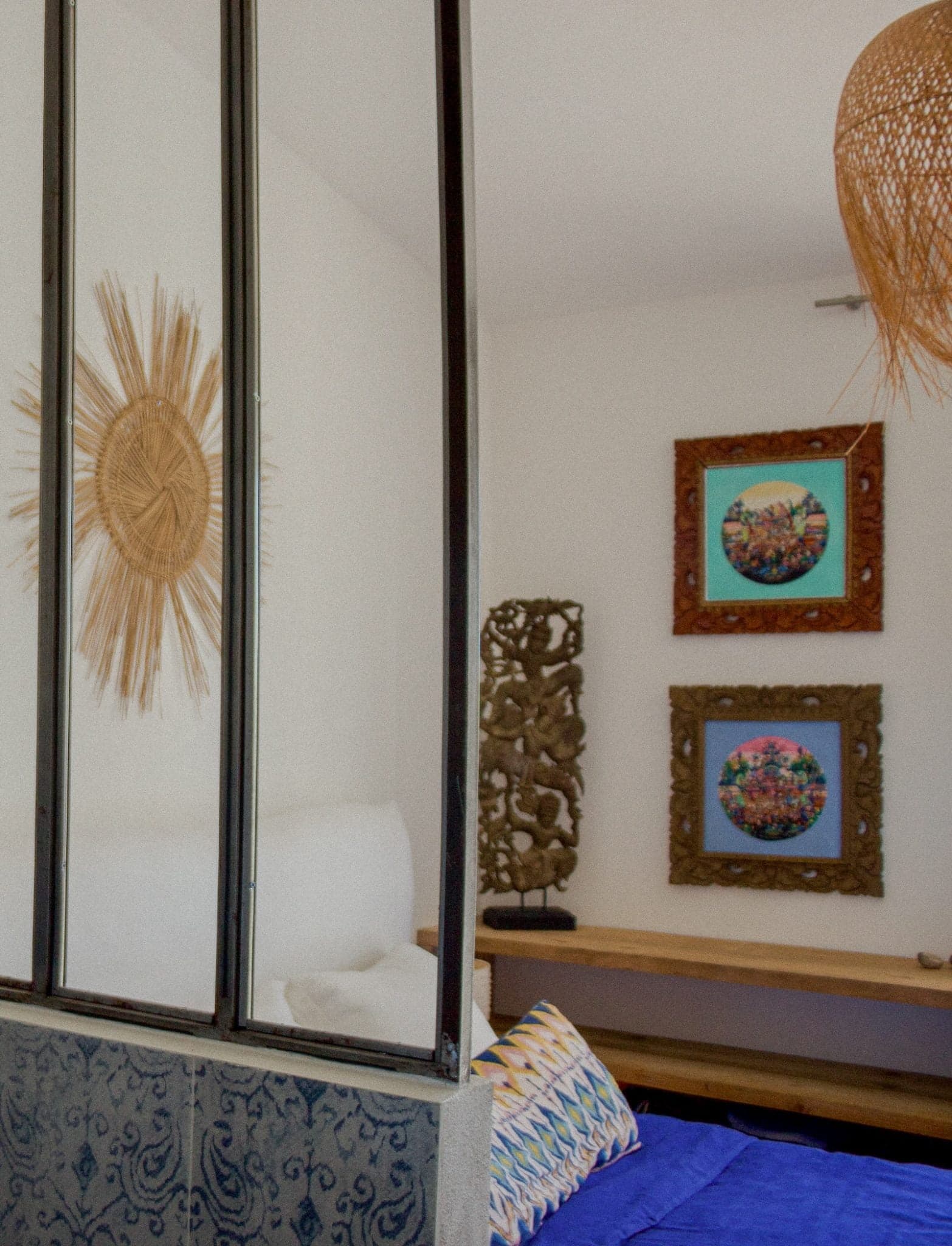 Une chambre douillette avec un couvre-lit bleu, des coussins décoratifs et une tête de lit blanche. Le mur est orné d'une décoration en forme de soleil tissé et de deux œuvres d'art colorées encadrées. Une sculpture en bois sculpté est placée sur une console. La pièce est divisée par une cloison en verre.