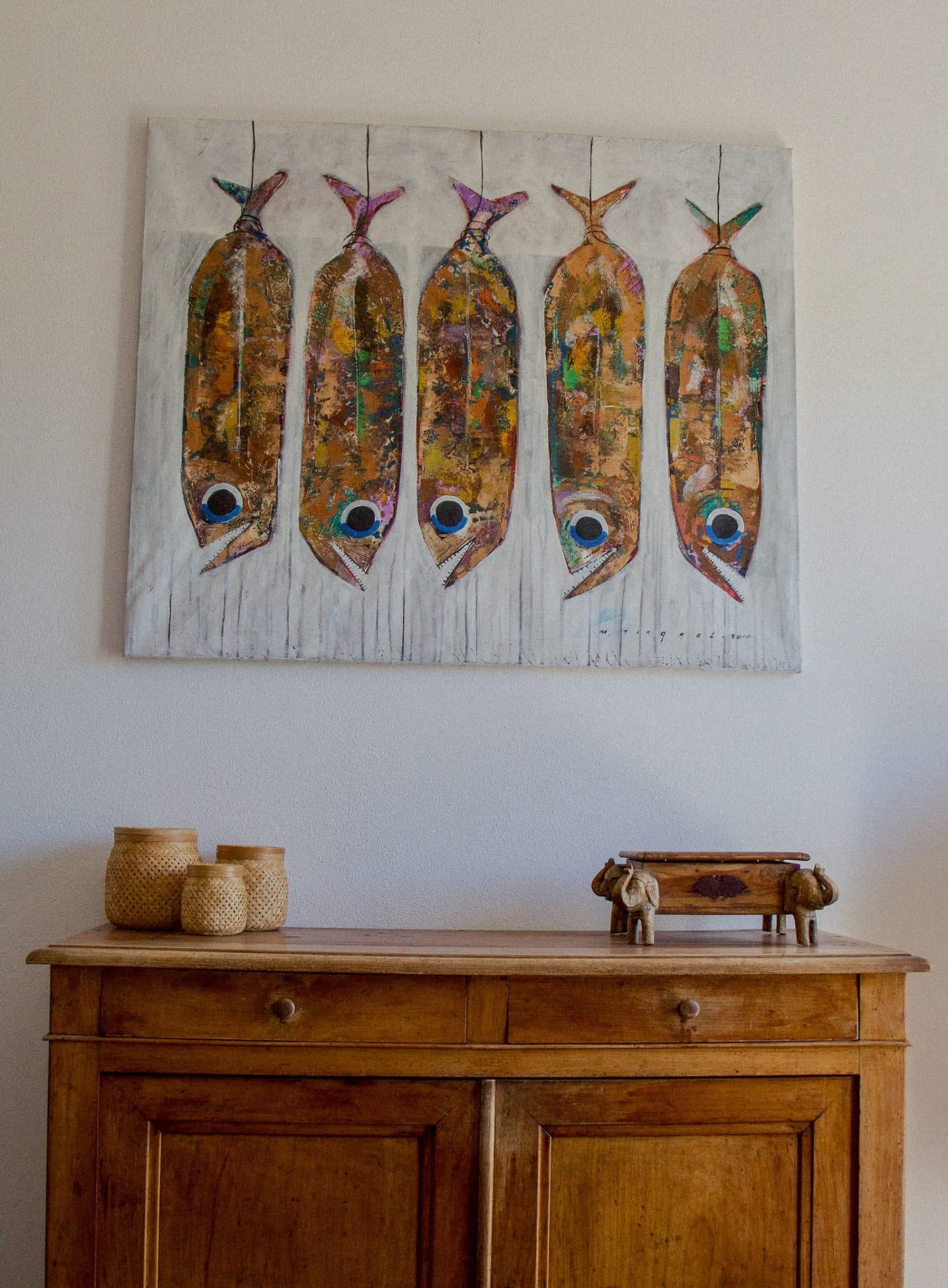Une commode en bois surmontée de paniers tressés et d'un petit objet décoratif se dresse contre un mur blanc. Au-dessus, un tableau représentant cinq poissons abstraits, aux couleurs vives et texturées et aux yeux proéminents, alignés verticalement avec leurs queues liées ensemble.