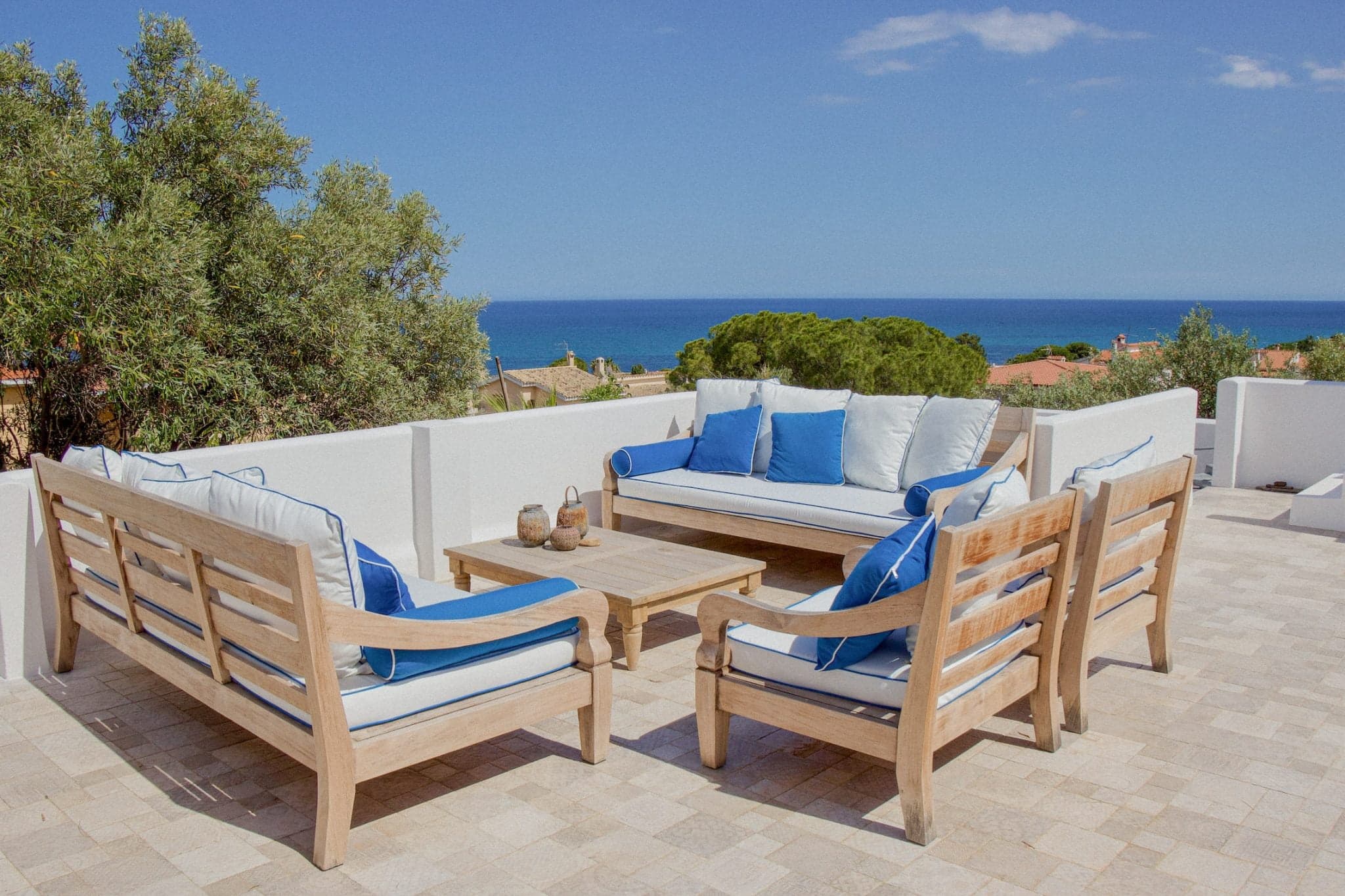 Un ensemble de patio avec des meubles en bois est disposé sur une terrasse extérieure carrelée. L'ensemble comprend deux canapés et deux fauteuils, tous avec des coussins blancs et des coussins décoratifs bleus. Une table basse en bois trône au centre. La terrasse donne sur une vue panoramique sur l'océan.