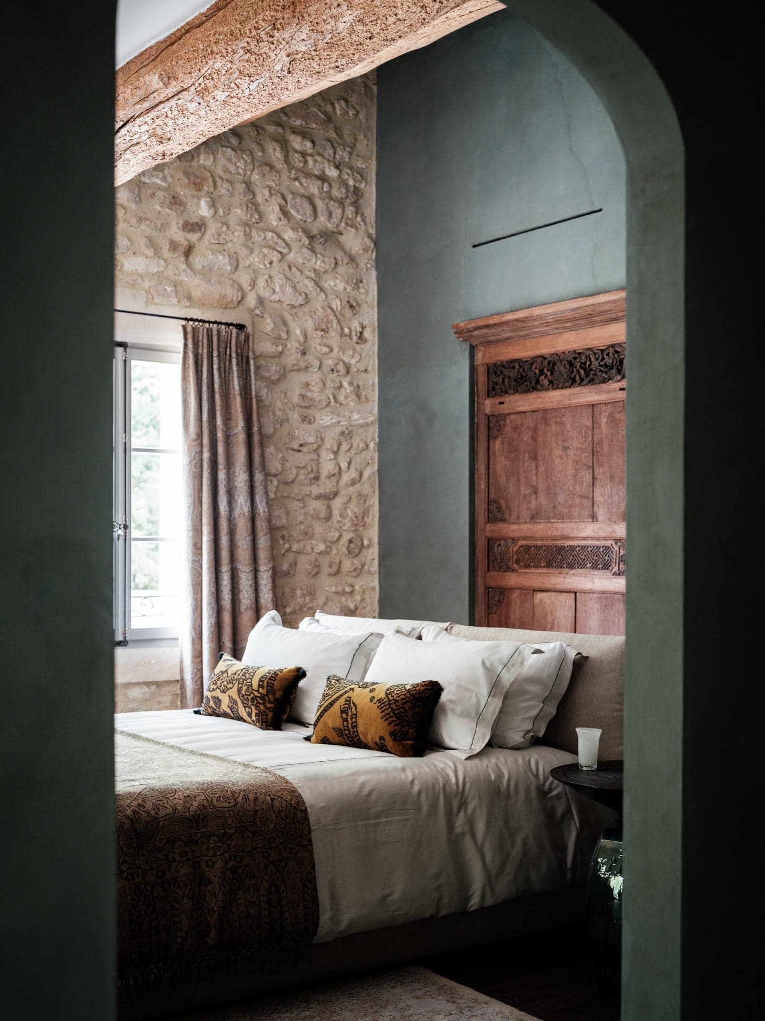 A cozy bedroom with stone walls and a vaulted entrance. The room features a wooden headboard, a neatly made bed with white and patterned linens, and decorative cushions in brown and mustard tones. A window draped with curtains provides soft natural light.