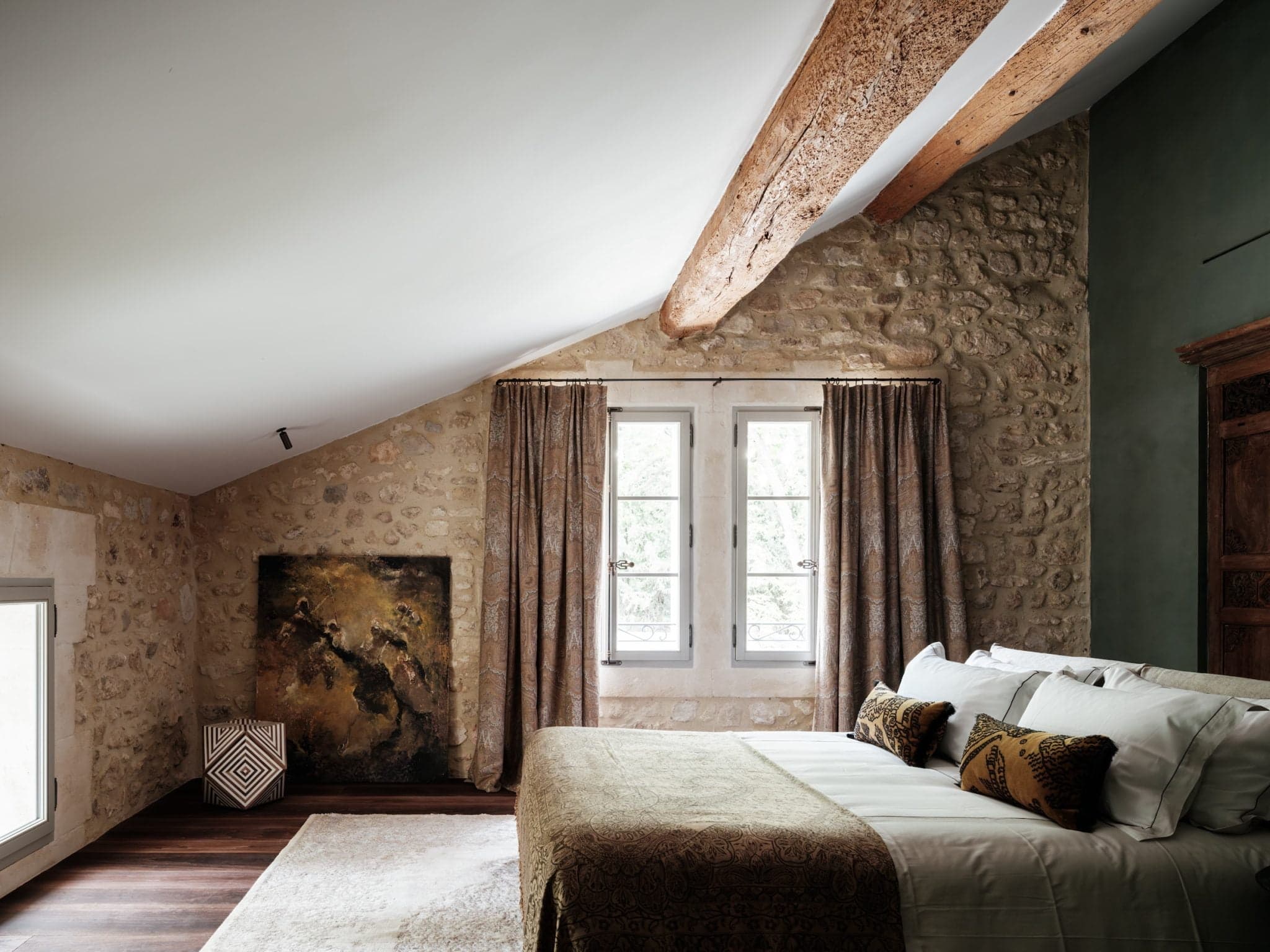 A cozy bedroom with rustic stone walls and exposed wooden beams. The room features a bed with white linens and decorative pillows, a large abstract painting resting on the floor, and two windows with beige curtains. The space is softly lit by natural light.