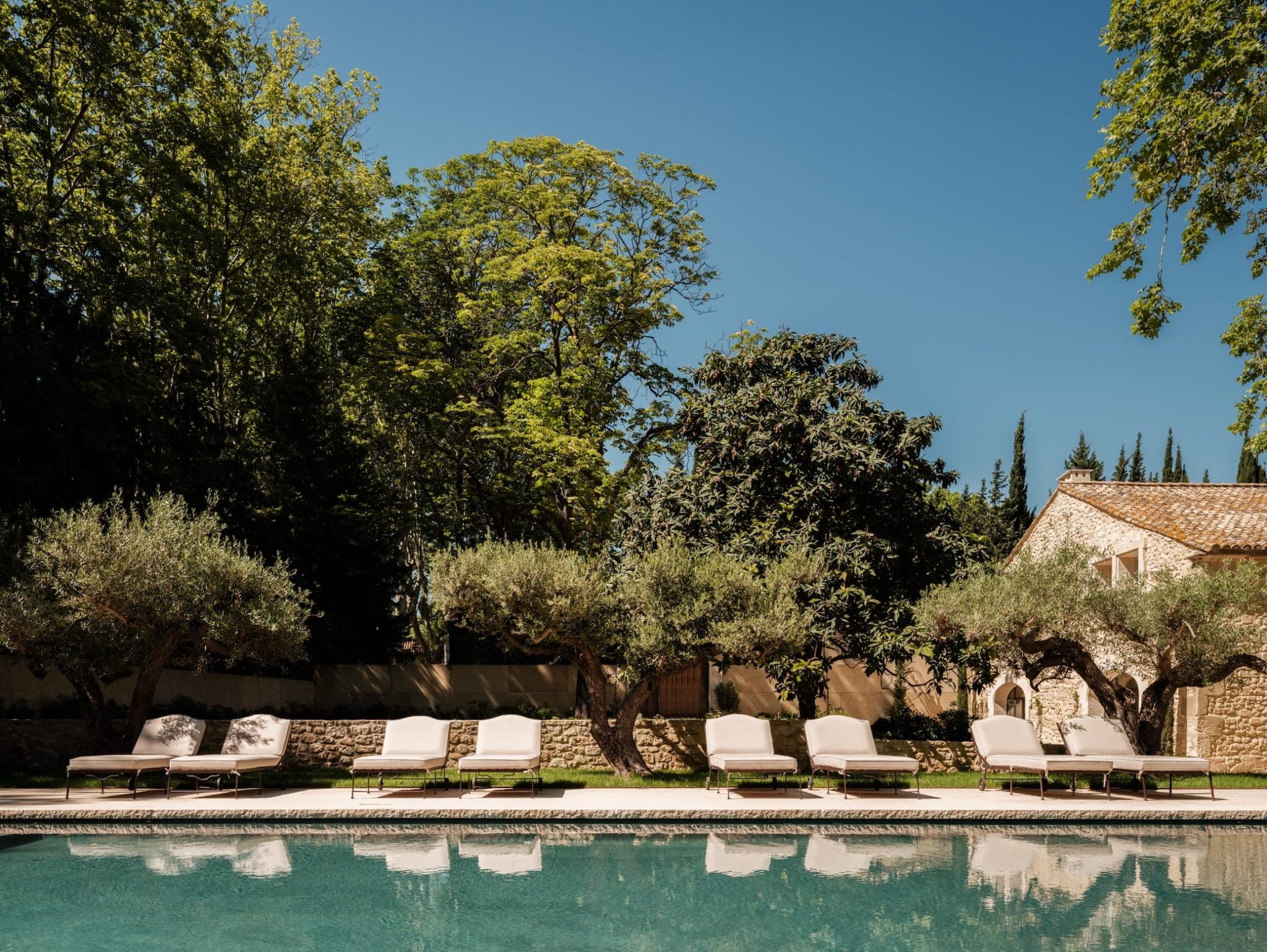 The image shows a serene outdoor pool with several lounge chairs arranged on the pool terrace, surrounded by greenery. Trees provide shade, and a rustic building with a tiled roof is in the background under a clear blue sky.