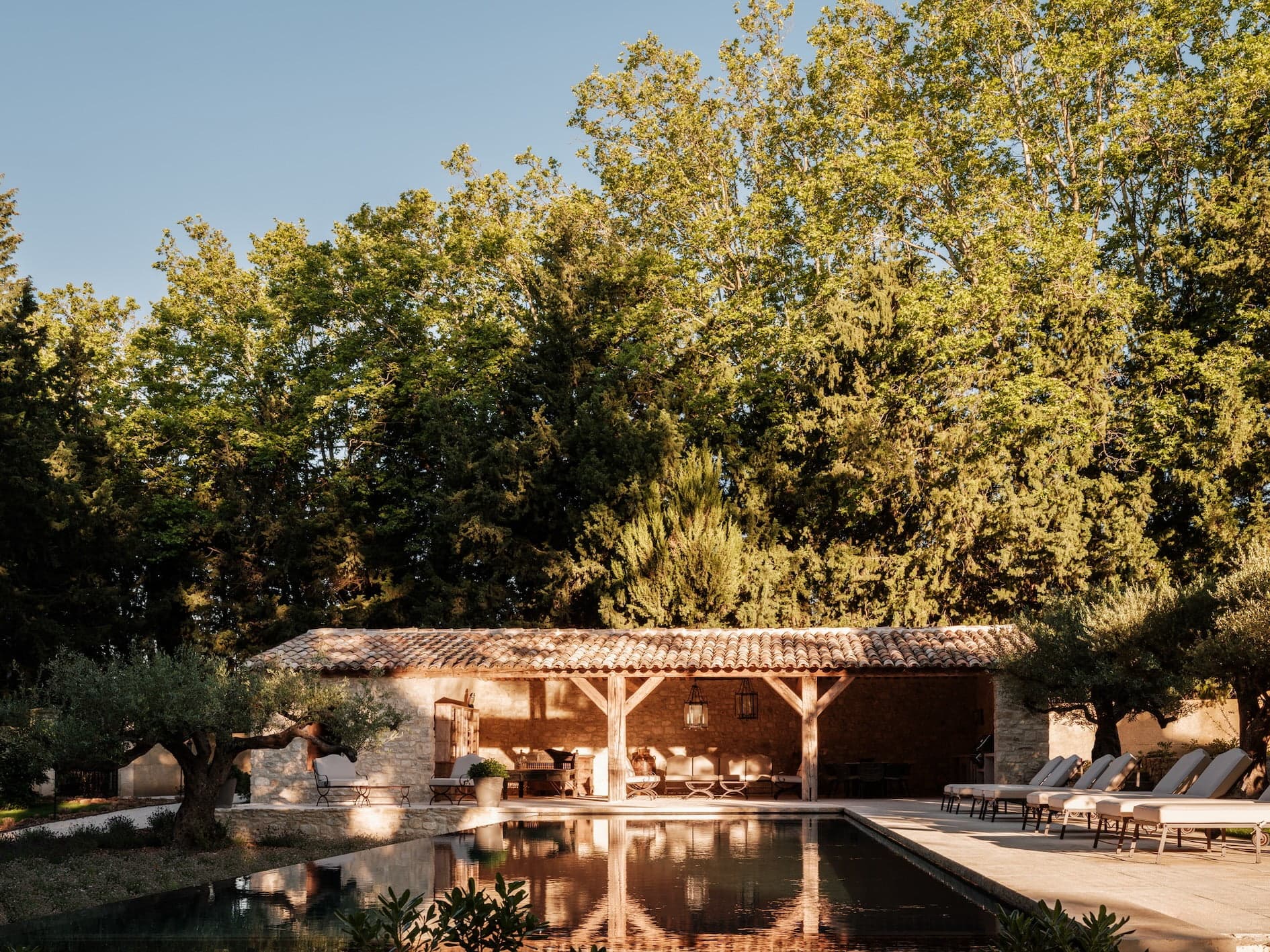 A serene outdoor pool area with a rustic stone building and a tiled roof. The building is shaded by tall trees, with comfortable lounge chairs lined up next to the pool. The water reflects the surrounding greenery, creating a tranquil atmosphere under a clear sky.