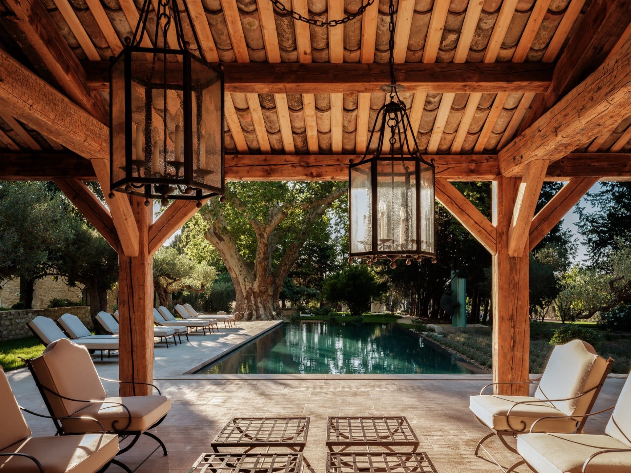 A covered outdoor space features wooden beams and large hanging lanterns. Comfortable chairs and tables are arranged under the structure, overlooking a tranquil pool surrounded by trees and greenery. Lounge chairs line the edge of the pool for relaxation.