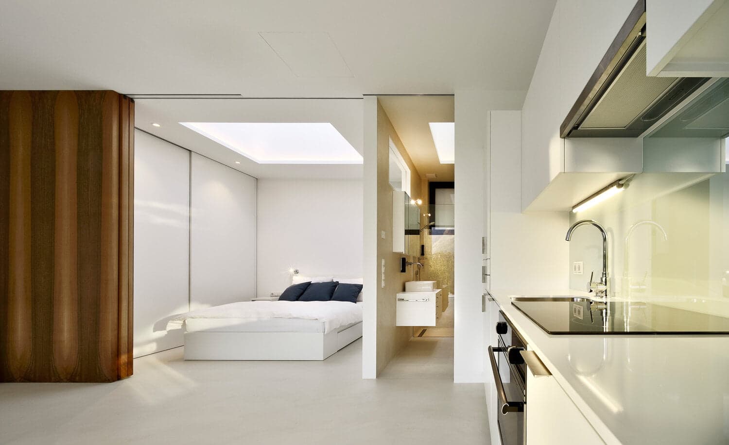 A modern, minimalist studio features a compact kitchen with sleek white cabinets and appliances on the right. On the left, a cozy bedroom with a white bed and blue pillows is separated by a sliding partition. A small bathroom with a sink and mirror is also visible.