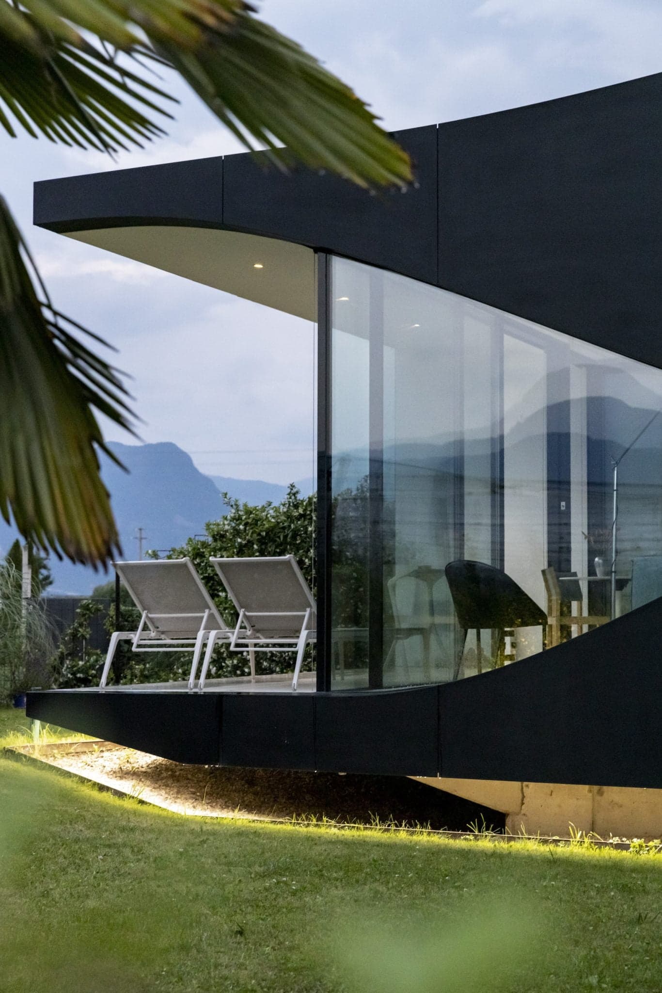 A modern building with black-edged features and large glass windows showcasing a stylish interior. Outside, two lounge chairs are positioned on a terrace. The background offers a panoramic view of the mountains under a cloudy sky, with a palm leaf visible in the foreground.