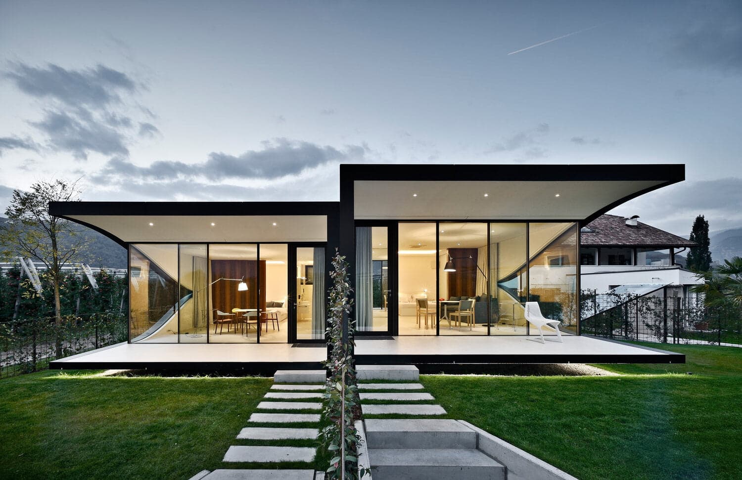 A modern house with large glass windows and a flat roof, showcasing an open interior. The exterior features a well-maintained lawn and a stone pathway leading to the entrance. The sky is partially cloudy with a hint of twilight light.