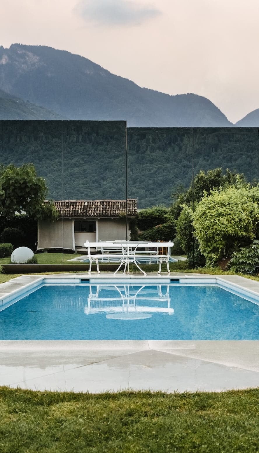 A serene outdoor pool in a landscaped courtyard, with a white table and chairs set nearby. Surrounding the pool are lush green bushes and a simple house. In the background, mountains under a cloudy sky create a tranquil atmosphere.