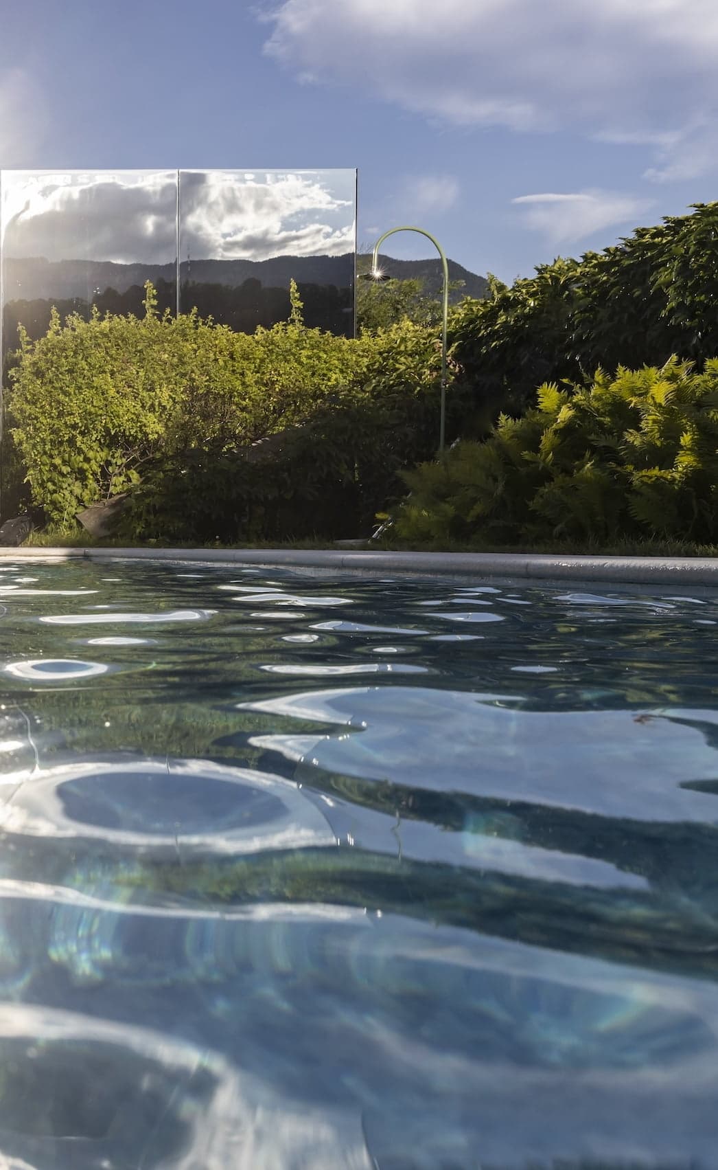 A serene outdoor pool with clear blue water reflects the surrounding greenery. Lush bushes and tall plants encircle the pool, while a mirrored wall seems to double the verdant landscape. Distant mountains and a partially cloudy sky are visible in the background.