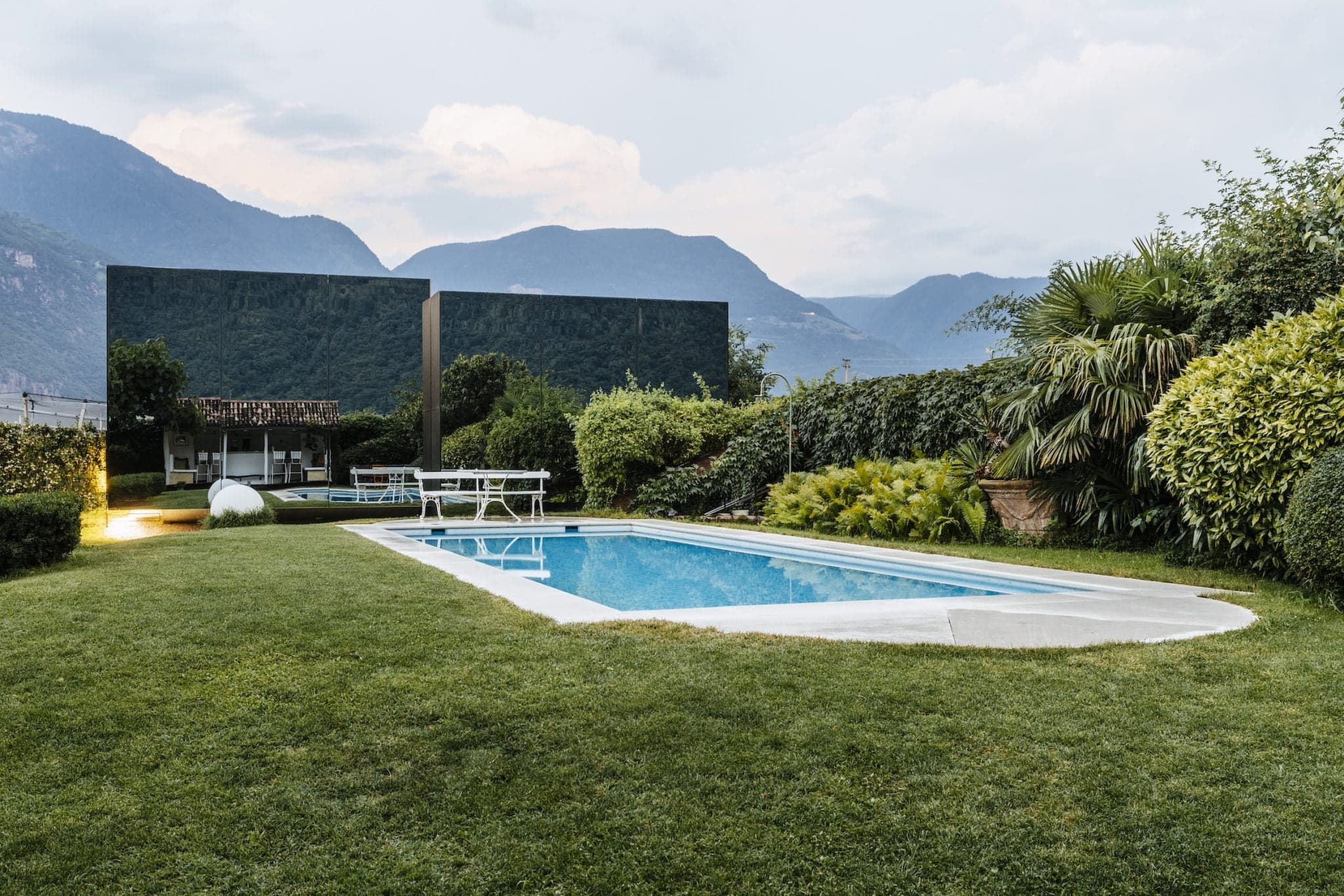 A serene backyard features a rectangular pool surrounded by lush green grass and various shrubs. In the background, two modern buildings with reflective glass facades blend into the mountainous landscape under a cloudy sky. A white table and chairs are positioned near the pool.