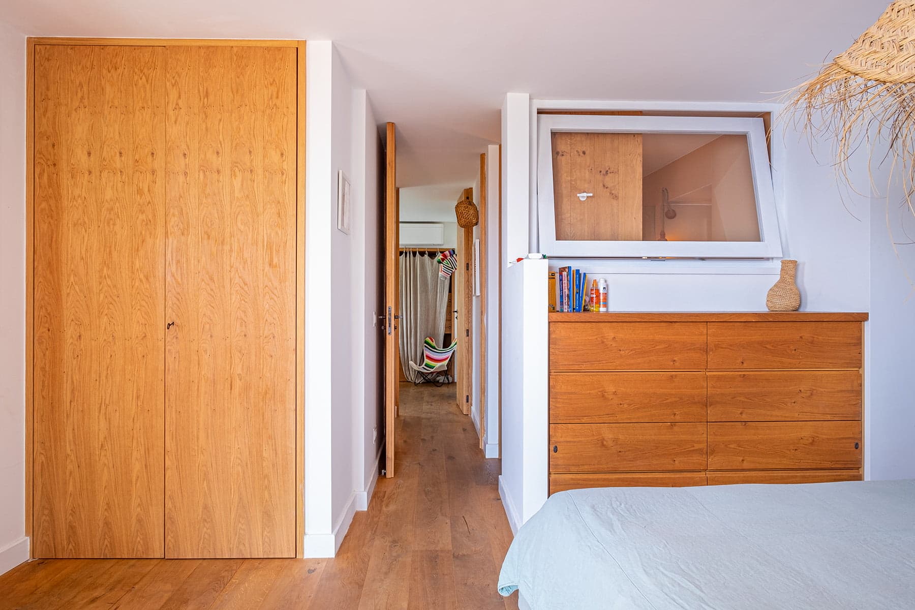 Une chambre lumineuse avec parquet et murs blancs comprend une grande armoire en bois à gauche et une grande commode en bois à droite. Une porte s'ouvre sur un couloir avec un hamac au bout. Une fenêtre au-dessus de la commode a une petite étagère avec des livres et des objets de décoration.