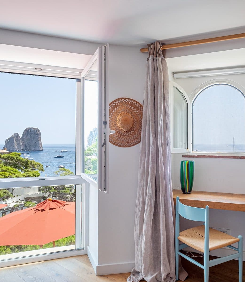 A cozy bedroom with an open window revealing a panoramic view of the ocean and rock formations. A desk with a blue chair and a green vase is located near an arched window. A straw hat is hung on the wall above light beige curtains. An orange umbrella is visible outside.