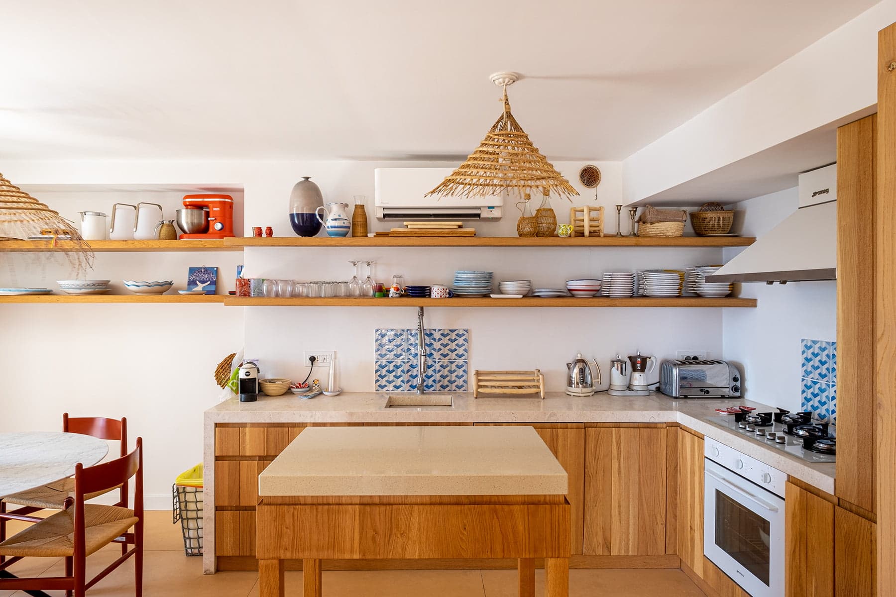 Une cuisine moderne avec des armoires en bois et des étagères ouvertes exposant divers plats, verres et ustensiles de cuisine. Une cuisinière et un four se trouvent à droite, et un îlot central se trouve au milieu. Deux suspensions tissées sont suspendues au plafond. Une chaise rouge se trouve à gauche.