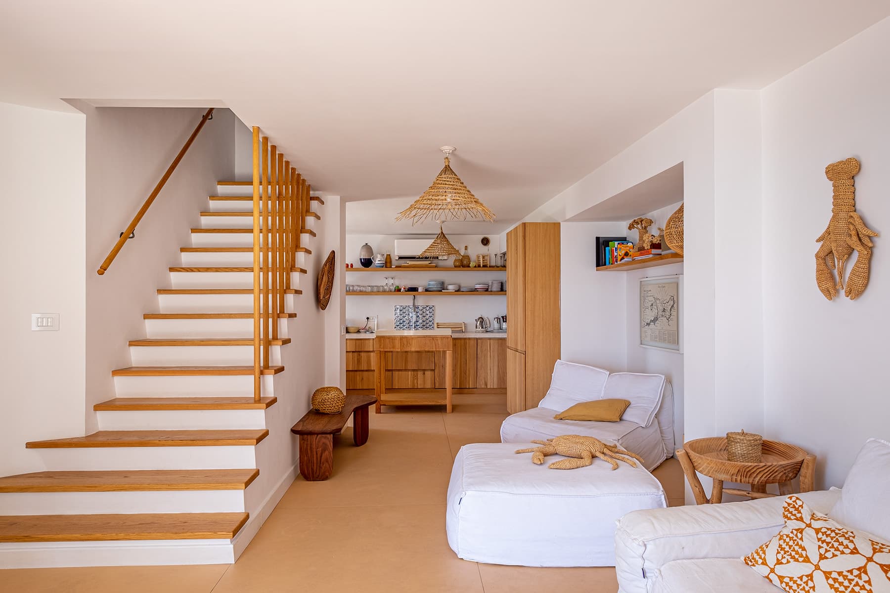A comfortable and well-lit living space with a wooden handrail staircase, white walls, and tiled flooring. The room includes upholstered white seating, wooden furniture, a small open kitchen, and beach-themed decor, such as wicker lampshades and hanging wall art.