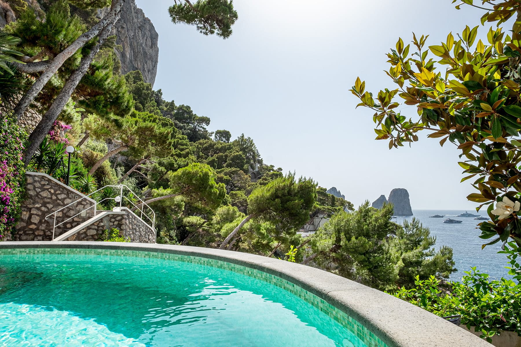 Panoramic view of a clear round pool overlooking a lush, green hill adorned with various trees and plants, with a rocky coastline and calm sea in the background under a clear sky.
