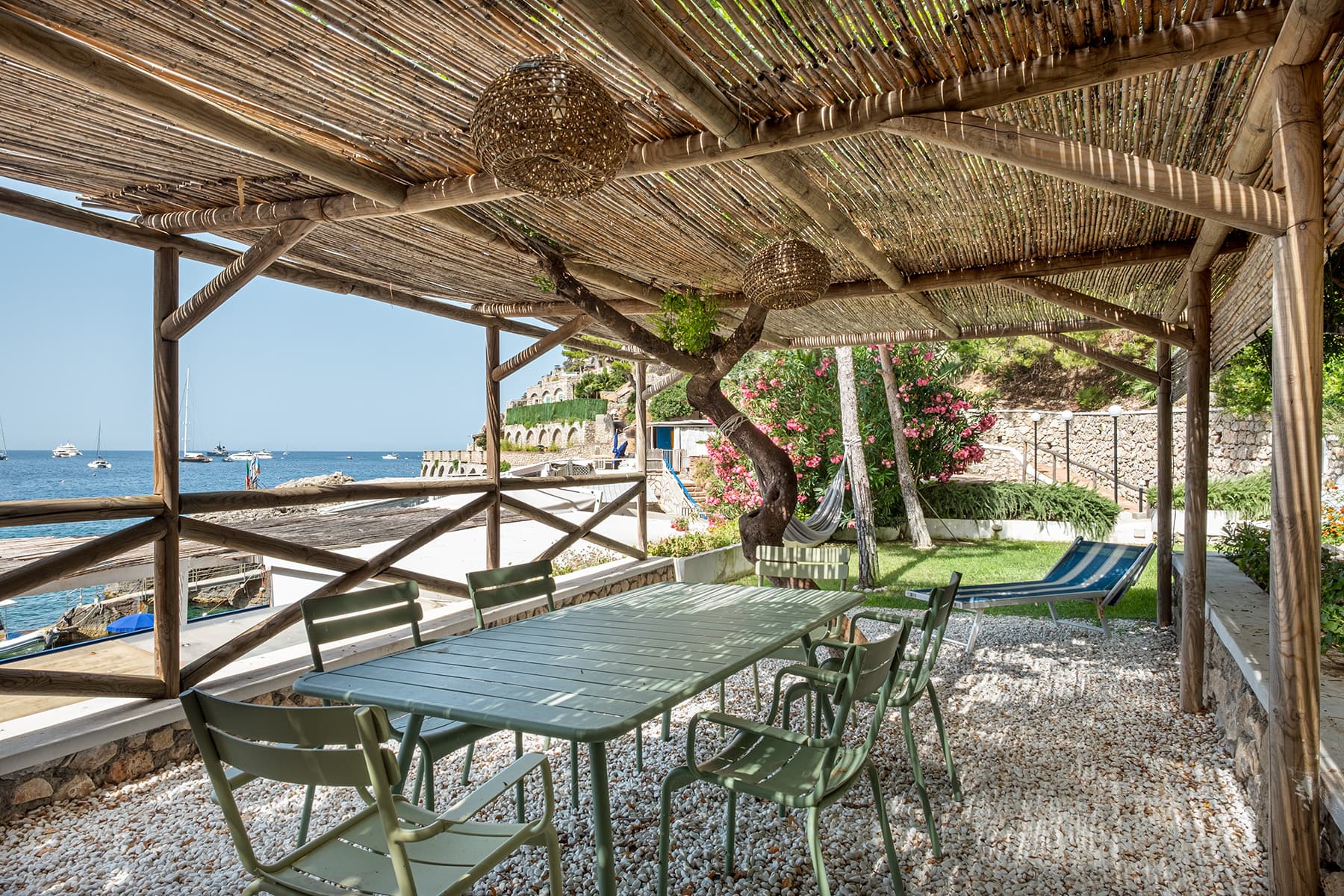 Un espace repas extérieur ombragé avec une table et des chaises vertes se trouve sur une terrasse en gravier donnant sur la mer. La pergola au-dessus est faite de poutres en bois et surmontée de bambou. En arrière-plan, il y a des bateaux sur l'eau et une végétation luxuriante avec des fleurs en fleurs.