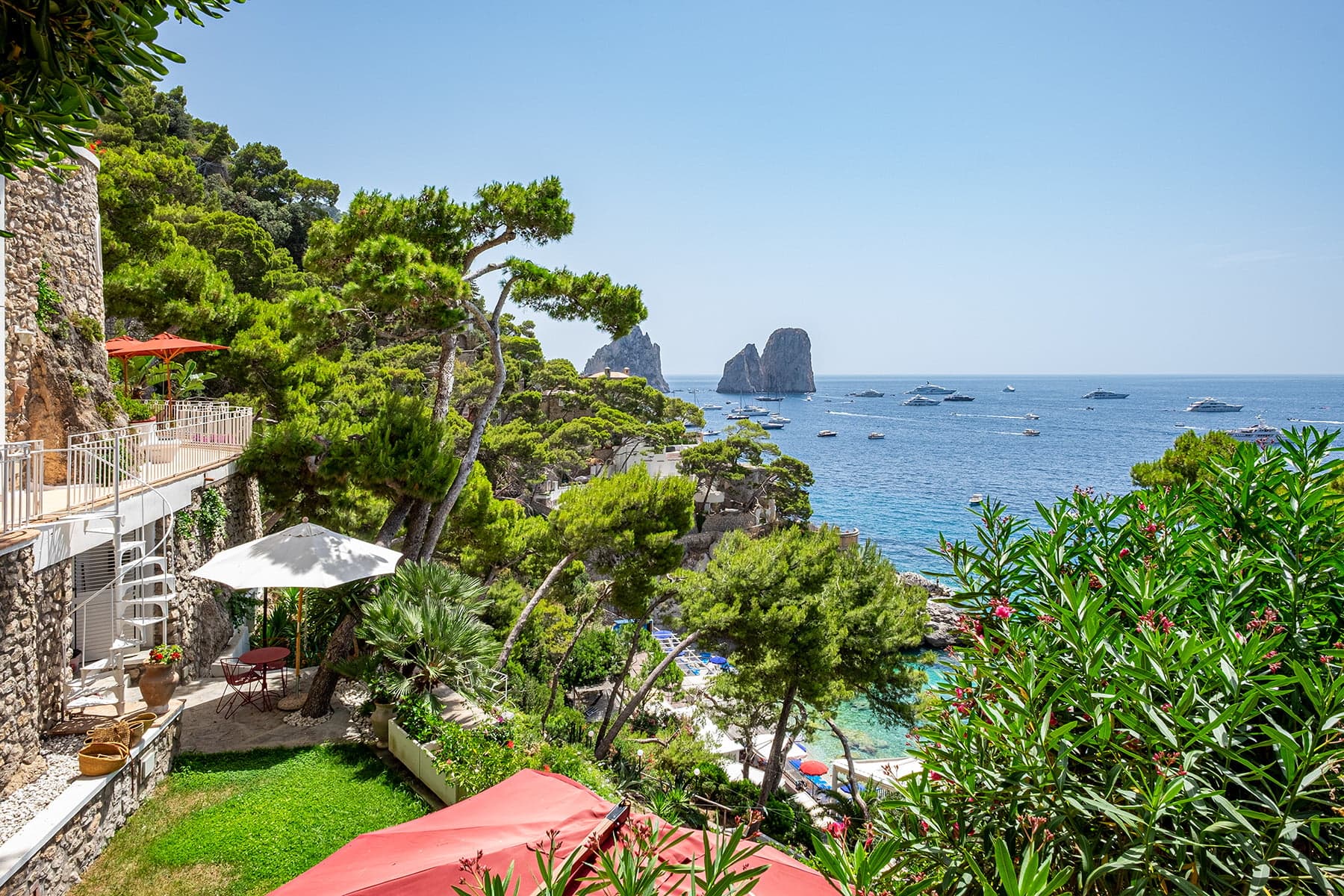 Vue panoramique d'une zone côtière avec une végétation verte luxuriante, des parasols rouges vibrants et un bâtiment sur la gauche. Des bateaux sont dispersés sur la mer bleue claire et des falaises rocheuses s'élèvent de l'eau en arrière-plan sous un ciel clair.
