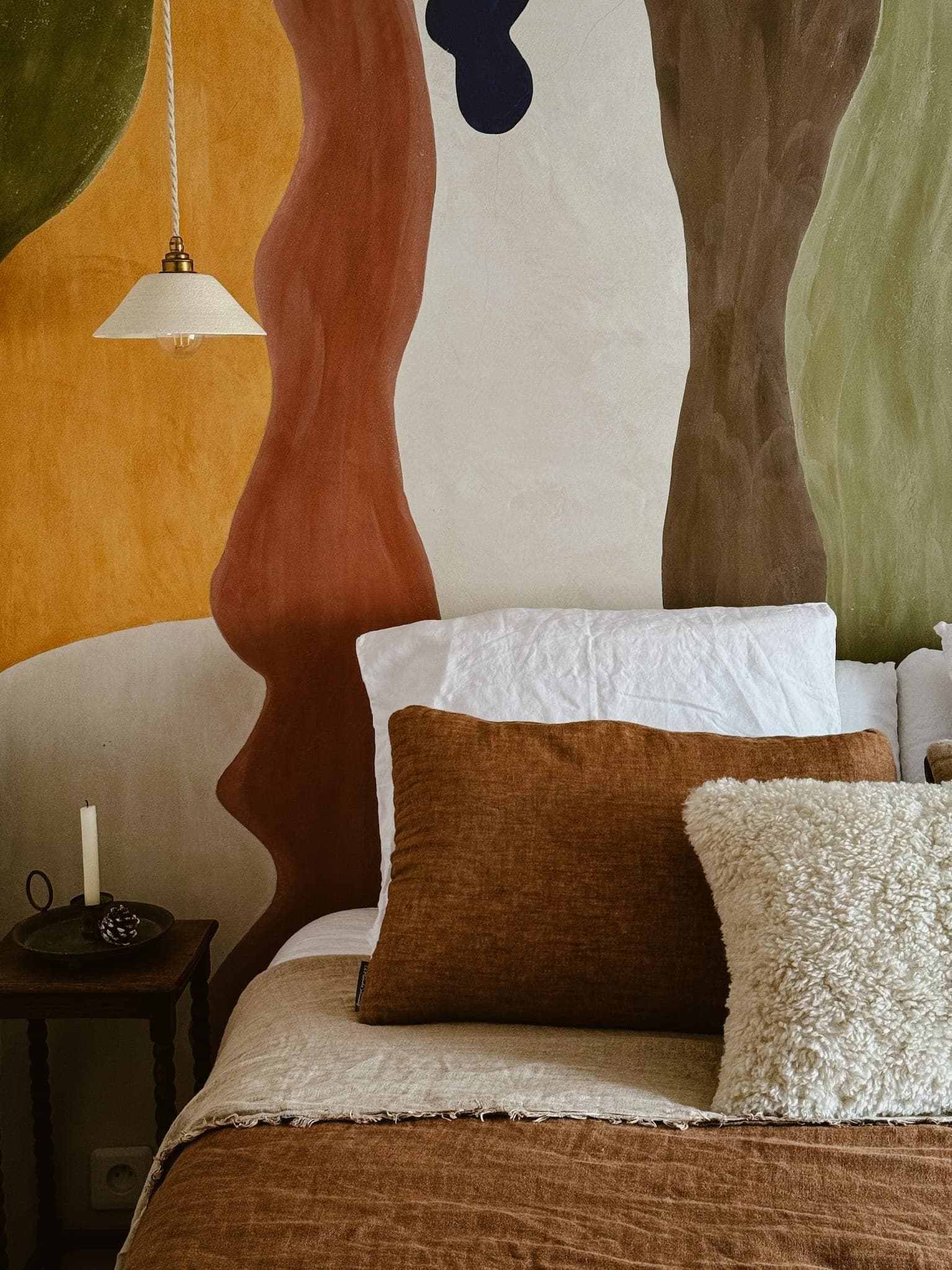 Une chambre confortable avec un lit avec des oreillers marron et crème et une couverture beige. Le mur du fond présente une fresque abstraite aux tons terreux de vert, orange, marron et bleu foncé. Une table d'appoint avec une bougie et une lampe suspendue se trouvent à côté du lit.