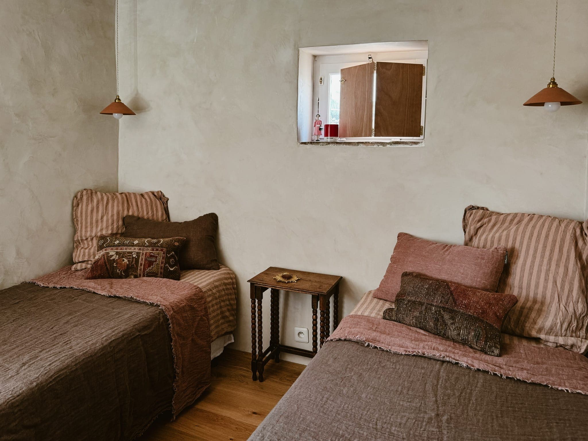 Une chambre confortable avec deux lits jumeaux et une literie marron à rayures. Une petite table en bois est placée entre les lits. Une petite fenêtre au-dessus de la table laisse entrer la lumière et des lampes suspendues sont suspendues au-dessus de chaque lit. La pièce dégage une atmosphère chaleureuse et rustique.