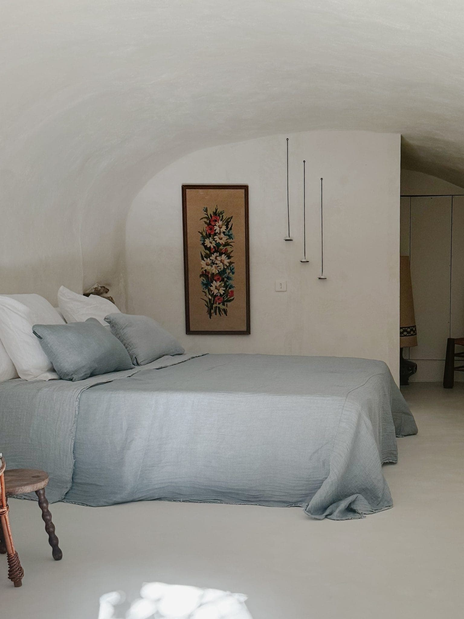 Une chambre minimaliste au plafond voûté comprend un lit recouvert de linge de lit bleu clair, un petit tabouret en bois et une œuvre d'art florale sur le mur. Trois suspensions sont suspendues au plafond, créant une atmosphère sereine et chaleureuse.