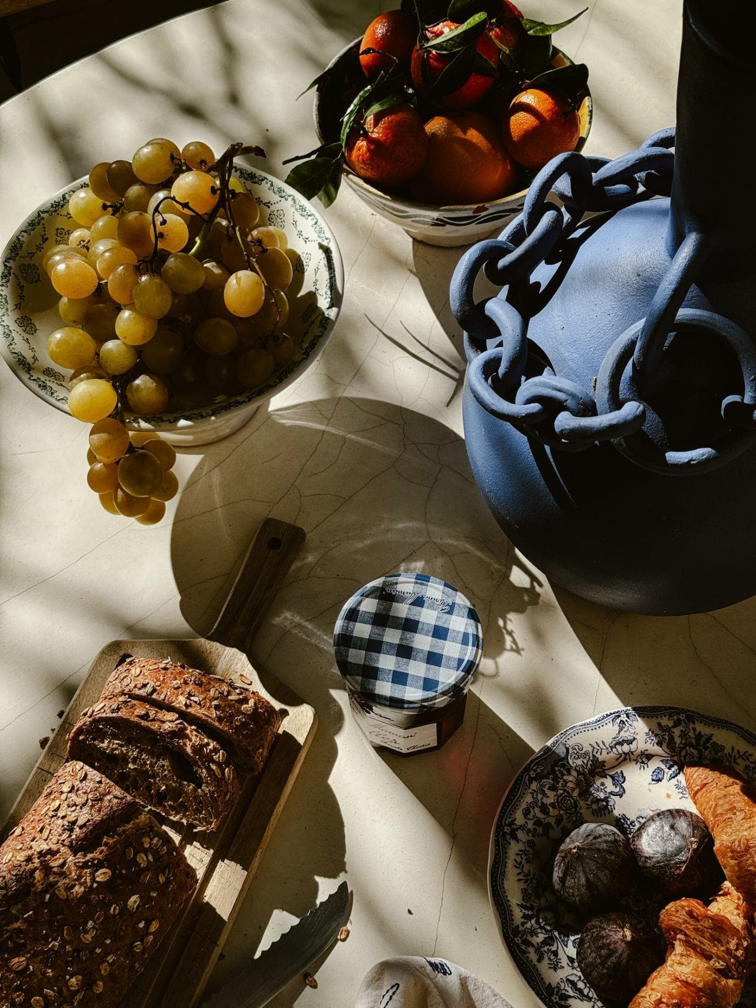 Une table ensoleillée avec du pain multigrains tranché sur une planche de bois, un pot de confiture, des figues violettes, des raisins dorés, un vase bleu avec un motif de chaîne et un bol d'oranges avec des feuilles. La scène est chaleureuse et rustique.
