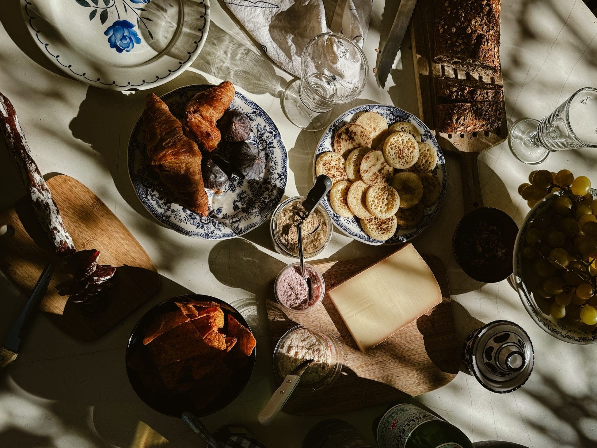 Une table ensoleillée avec une variété d'aliments : des croissants, des crumpets, du pain en tranches, des raisins, un bloc de fromage, des tranches de saucisse, un petit bol de trempette et une canette de boisson. Des assiettes, des verres et des ombres ajoutent à l'esthétique rustique.