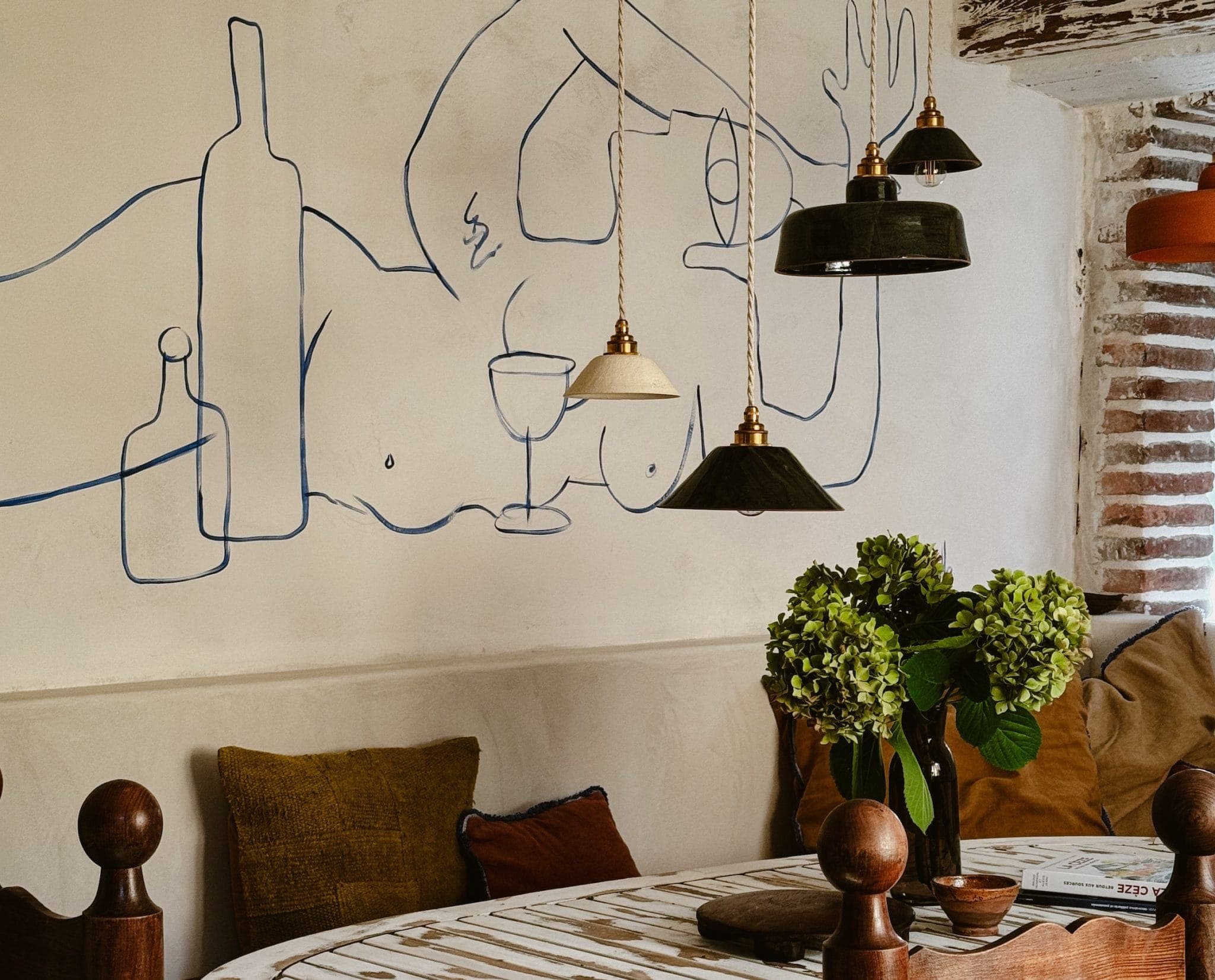 Salle à manger confortable avec une table ronde, des chaises en bois et des hortensias verts. Des suspensions diffusent une lumière chaleureuse. Le mur présente un dessin au trait minimaliste d'une figure allongée avec des bouteilles de vin et un verre. Détails rustiques en brique et en bois.
