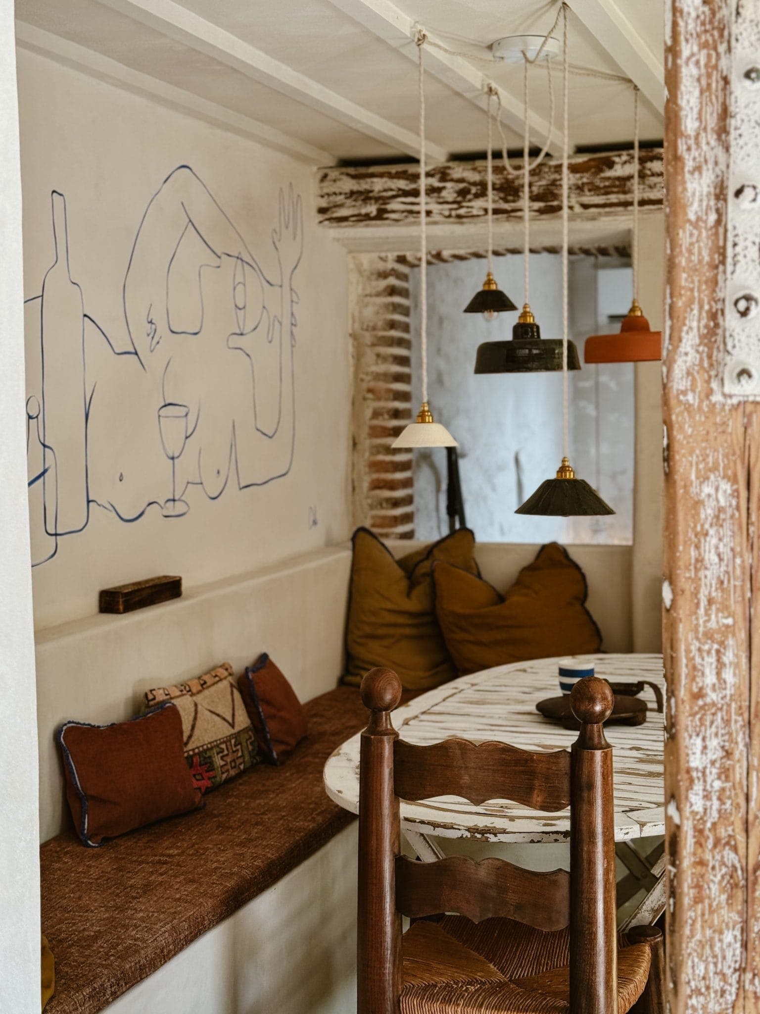 Cozy dining nook with a rustic wooden table, surrounded by cushioned benches. Pendant lights hang above, and a minimalist line drawing of a reclining figure with a wine bottle decorates the wall. Earthy tones create a warm atmosphere.