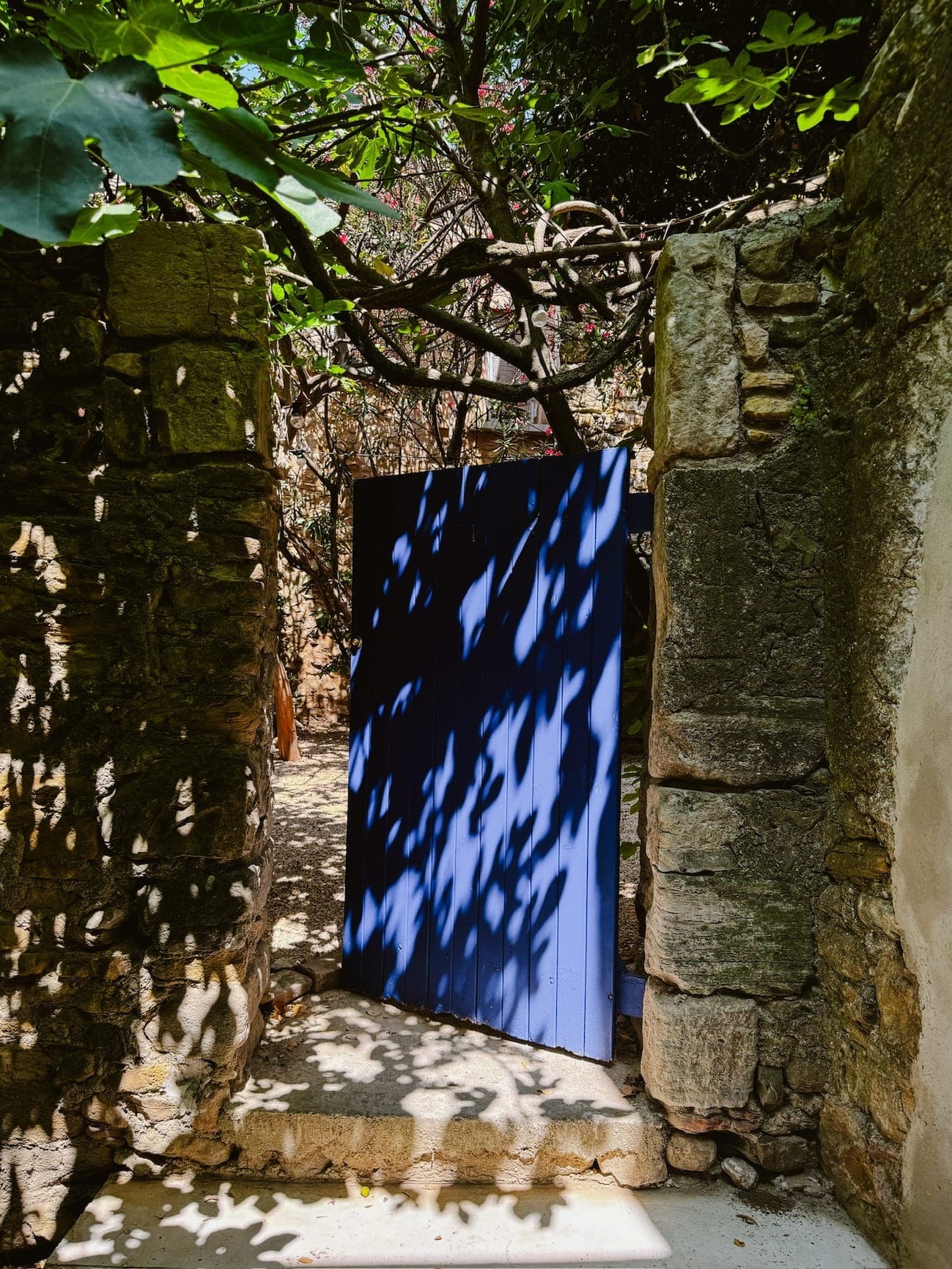 Une arche en pierre rustique mène à une porte d'un bleu éclatant, partiellement ouverte. La lumière du soleil filtre à travers les arbres au-dessus, projetant des ombres complexes sur le chemin et la porte. Une végétation luxuriante entoure la scène, créant une atmosphère sereine et accueillante.