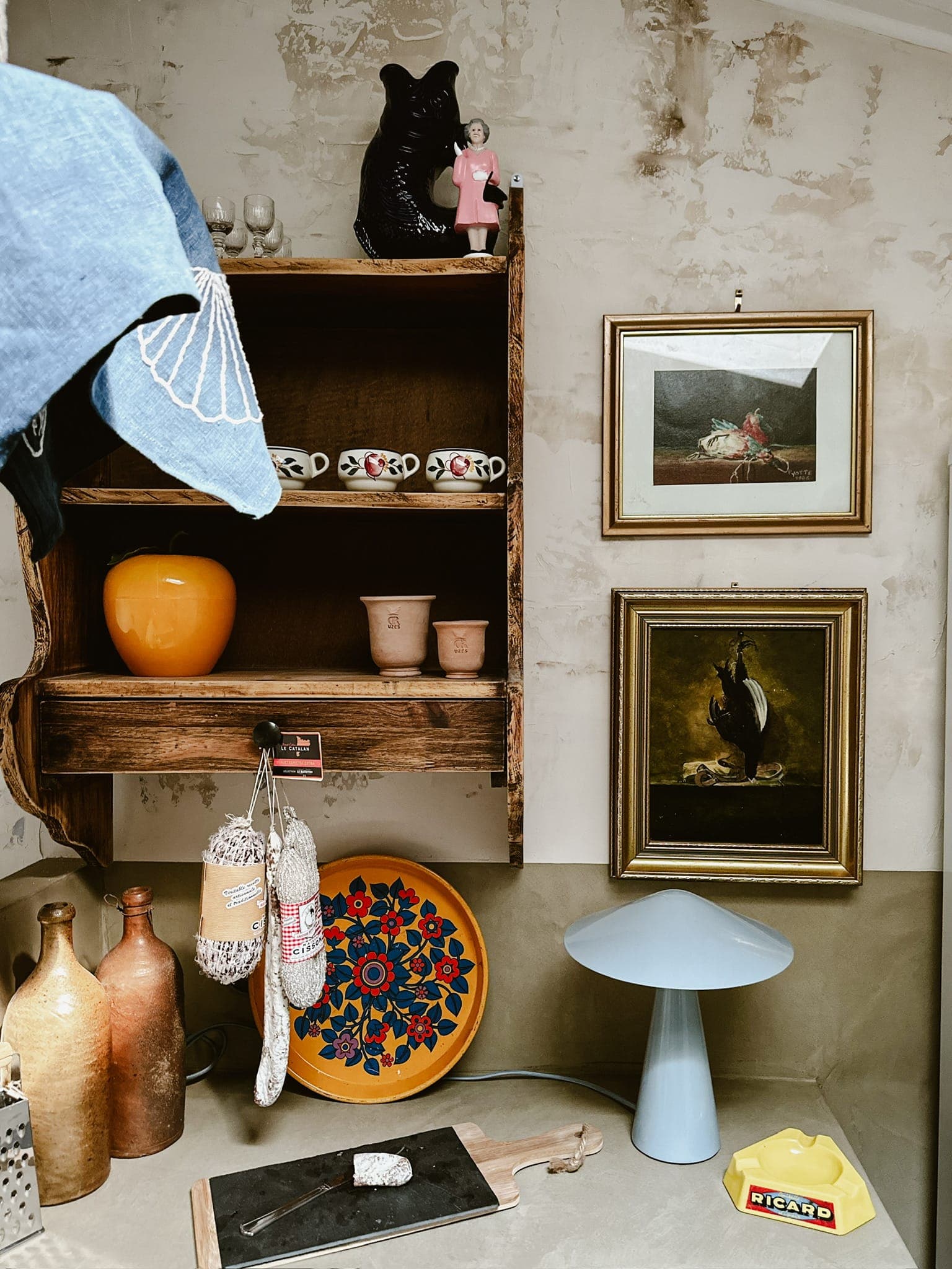 Un coin cuisine rustique avec une étagère en bois sur laquelle sont posés des tasses et des petits pots, entourés d'œuvres d'art encadrées. Une assiette colorée, une lampe bleue et des objets décoratifs comme des cruches en céramique et une figurine de chat noir complètent la scène.