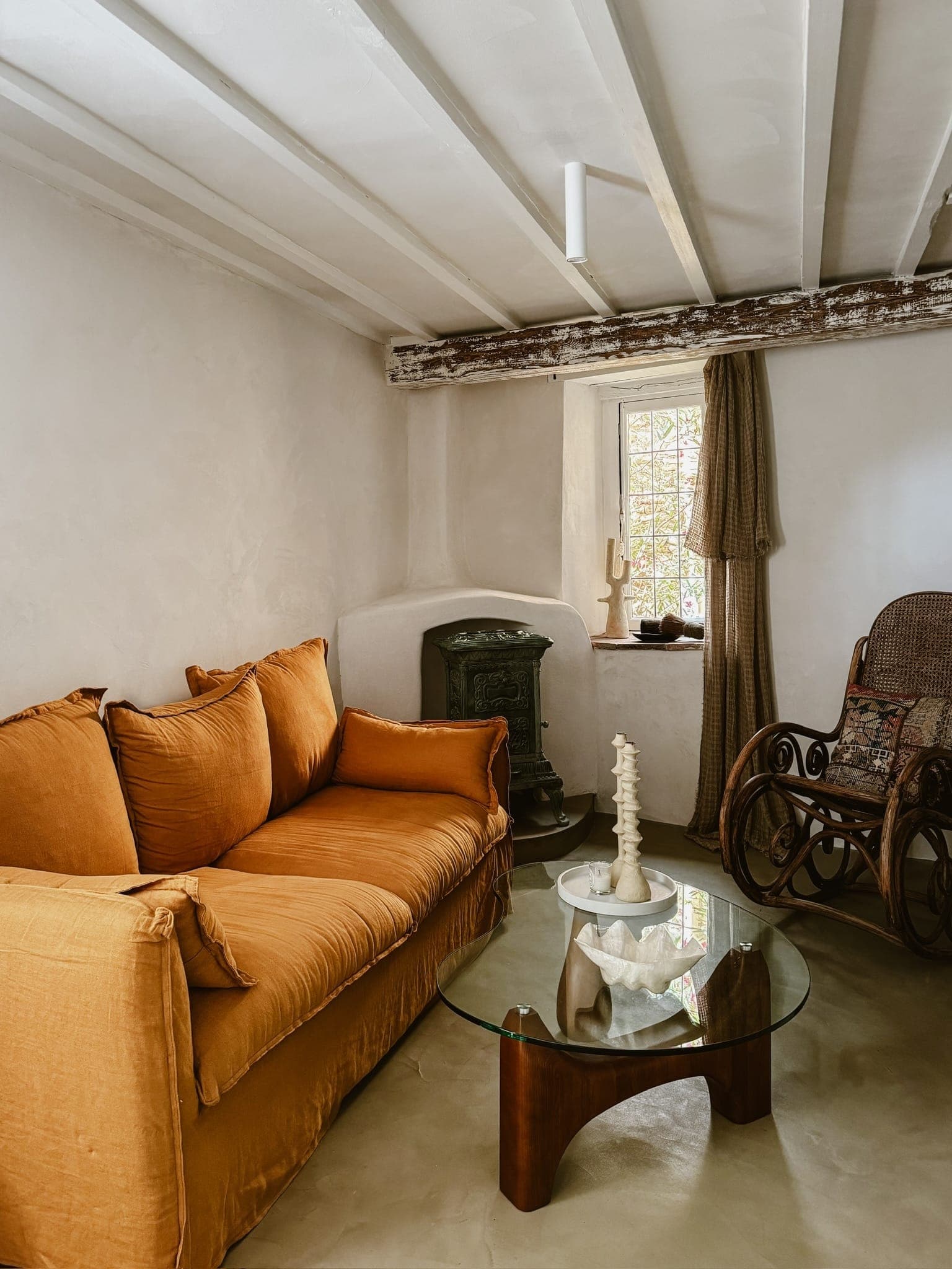 Une pièce chaleureuse avec un canapé orange, une table basse en verre avec des objets décoratifs et une chaise à bascule. Un plafond rustique à poutres en bois et une petite fenêtre avec un rideau ajoutent du charme. Un poêle à bois se trouve dans le coin.