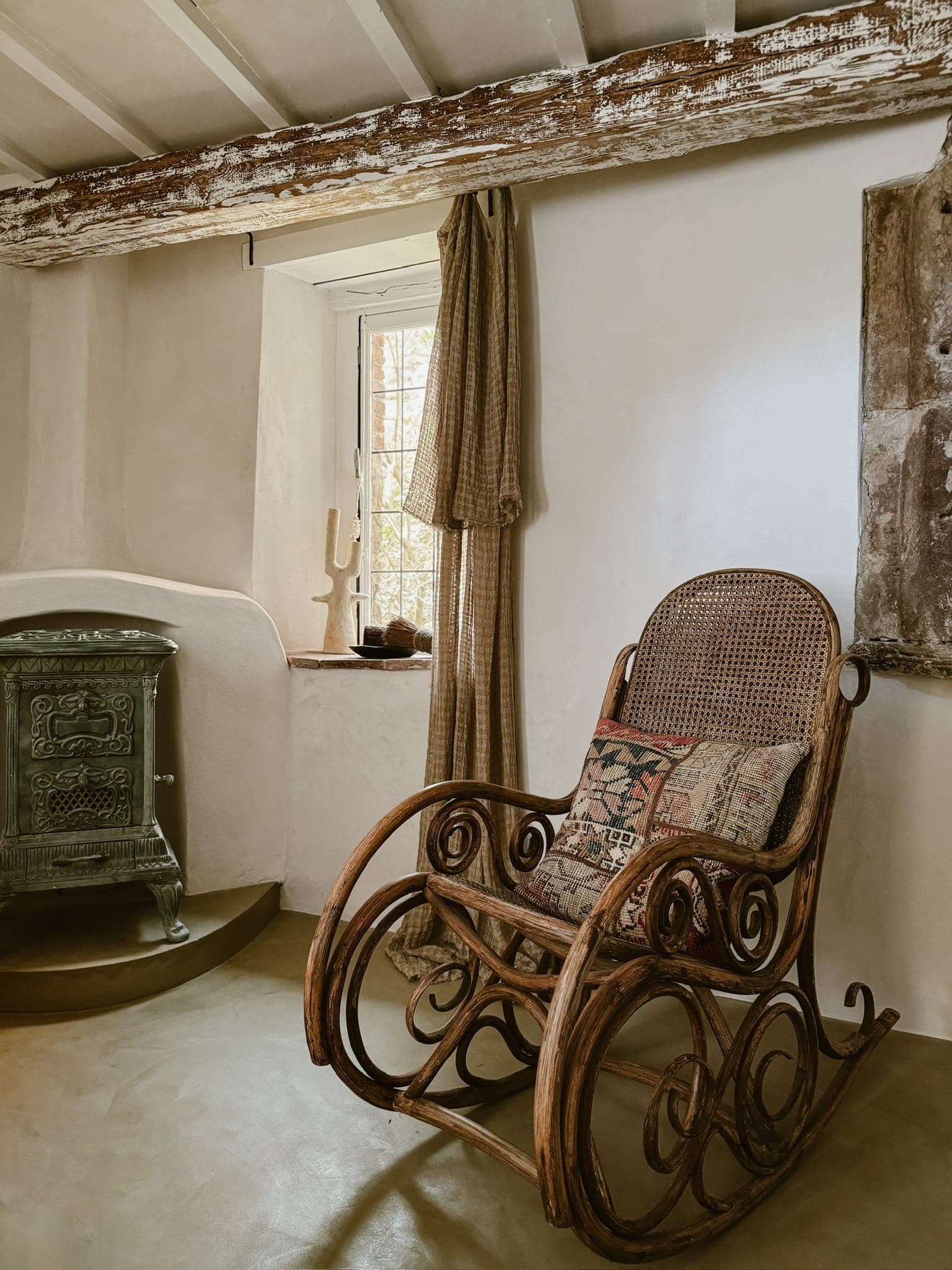 Une pièce rustique est dotée d'un fauteuil à bascule vintage avec un coussin à motifs. À côté, un poêle en fonte vert se dresse sur une plate-forme surélevée. Une fenêtre avec des rideaux à carreaux permet à la lumière naturelle d'illuminer doucement l'espace, mettant en valeur les poutres en bois au-dessus.